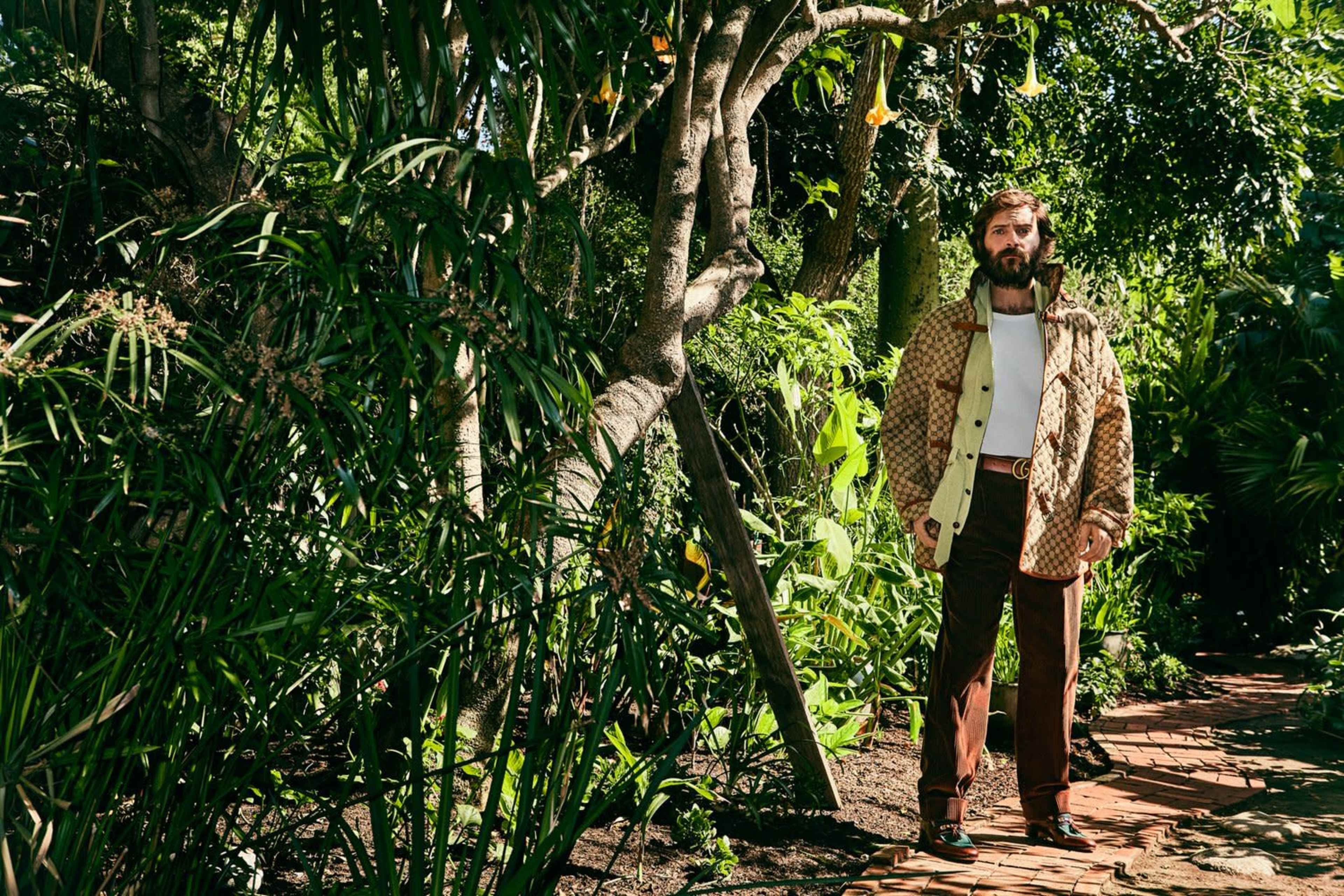A man dressed in a patterned jacket and brown trousers stands among lush greenery in a garden.