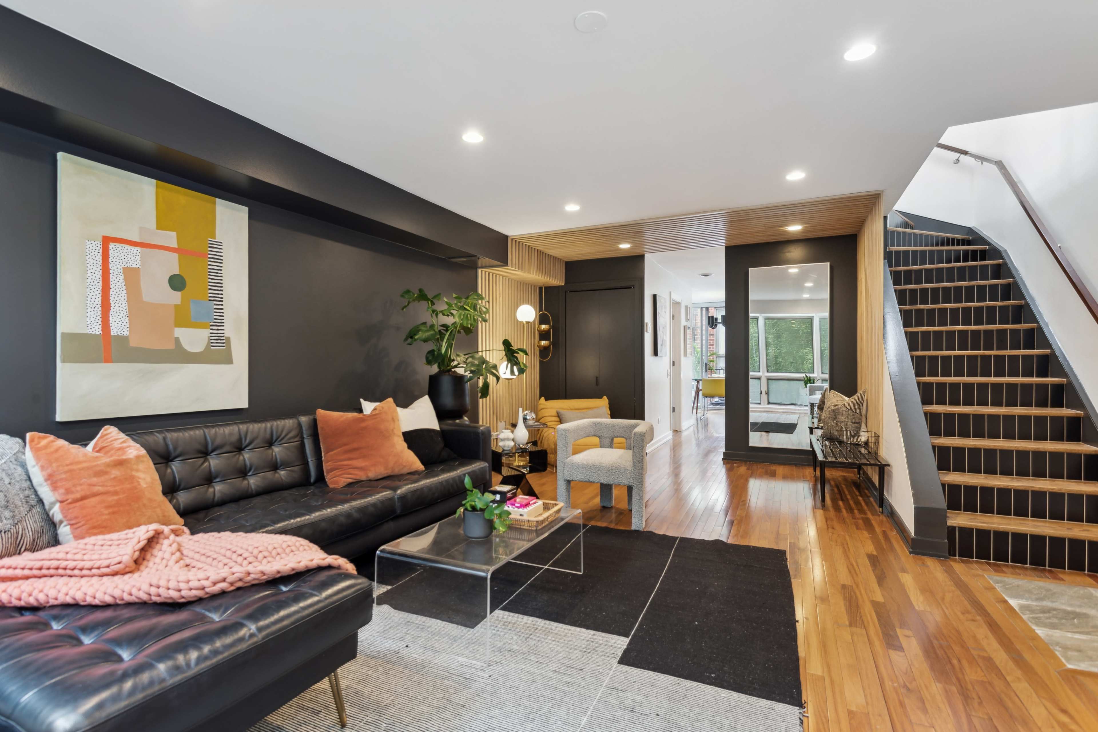 A modern living room featuring a black leather sofa with colorful pillows, a glass coffee table, and a staircase leading to an upper level.