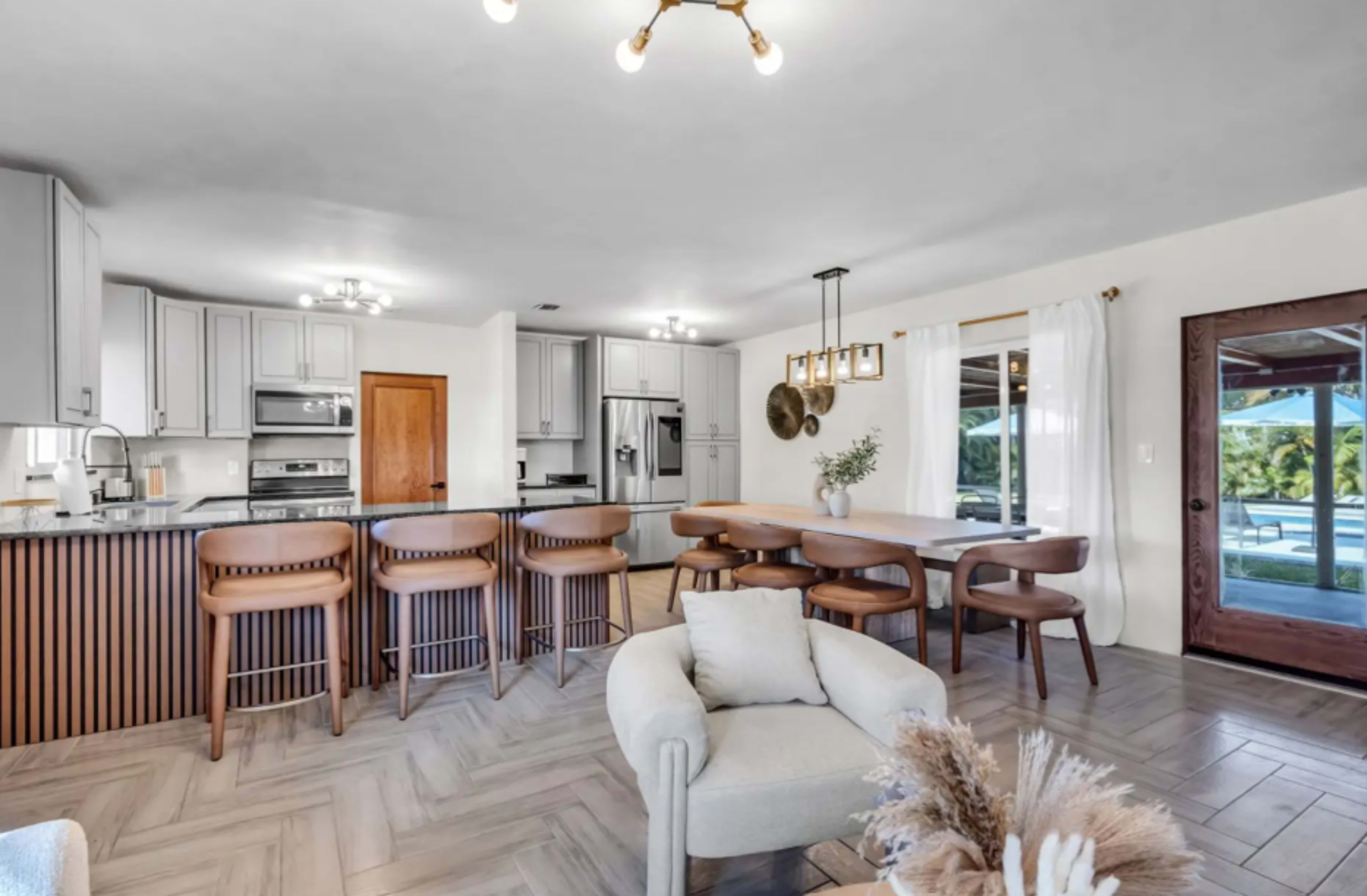 The image shows a modern kitchen and dining area with a central island, wooden dining table, and comfortable seating.