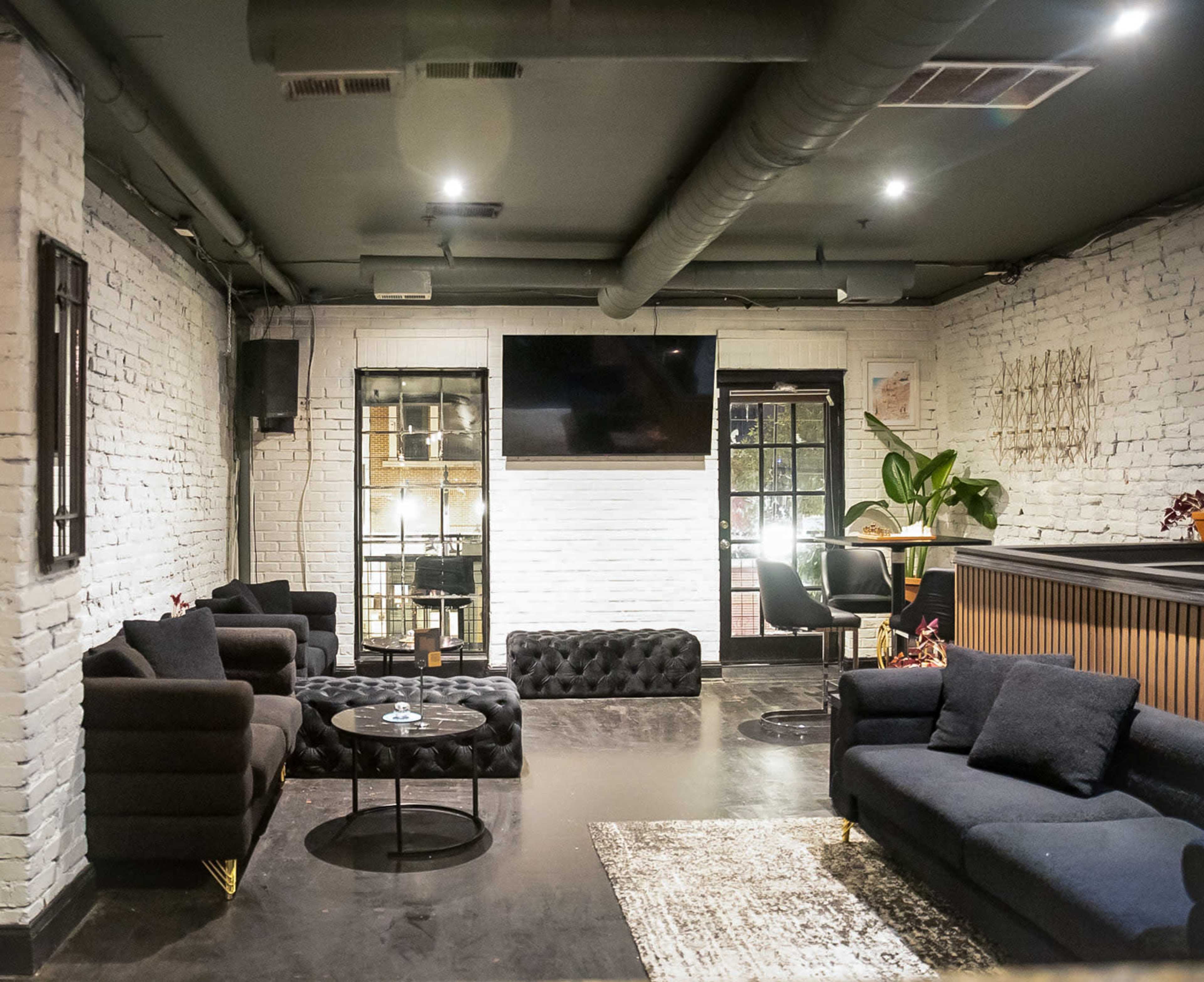 The image shows a modern lounge area with dark sofas, a large television mounted on a white brick wall, and a contrasting black and white color scheme.