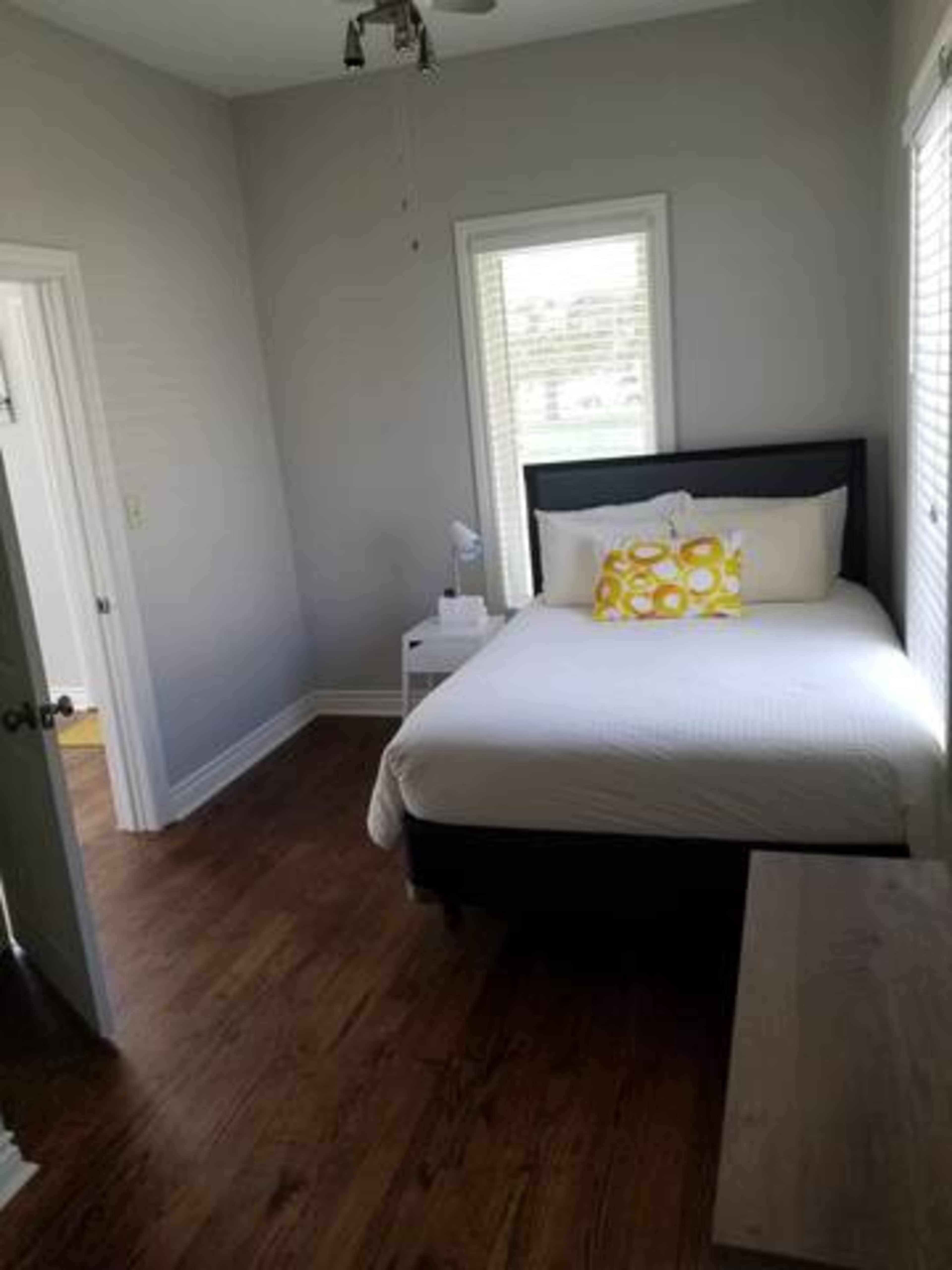 A small bedroom with a bed adorned with a yellow pillow, a nightstand, and natural light coming through two windows.