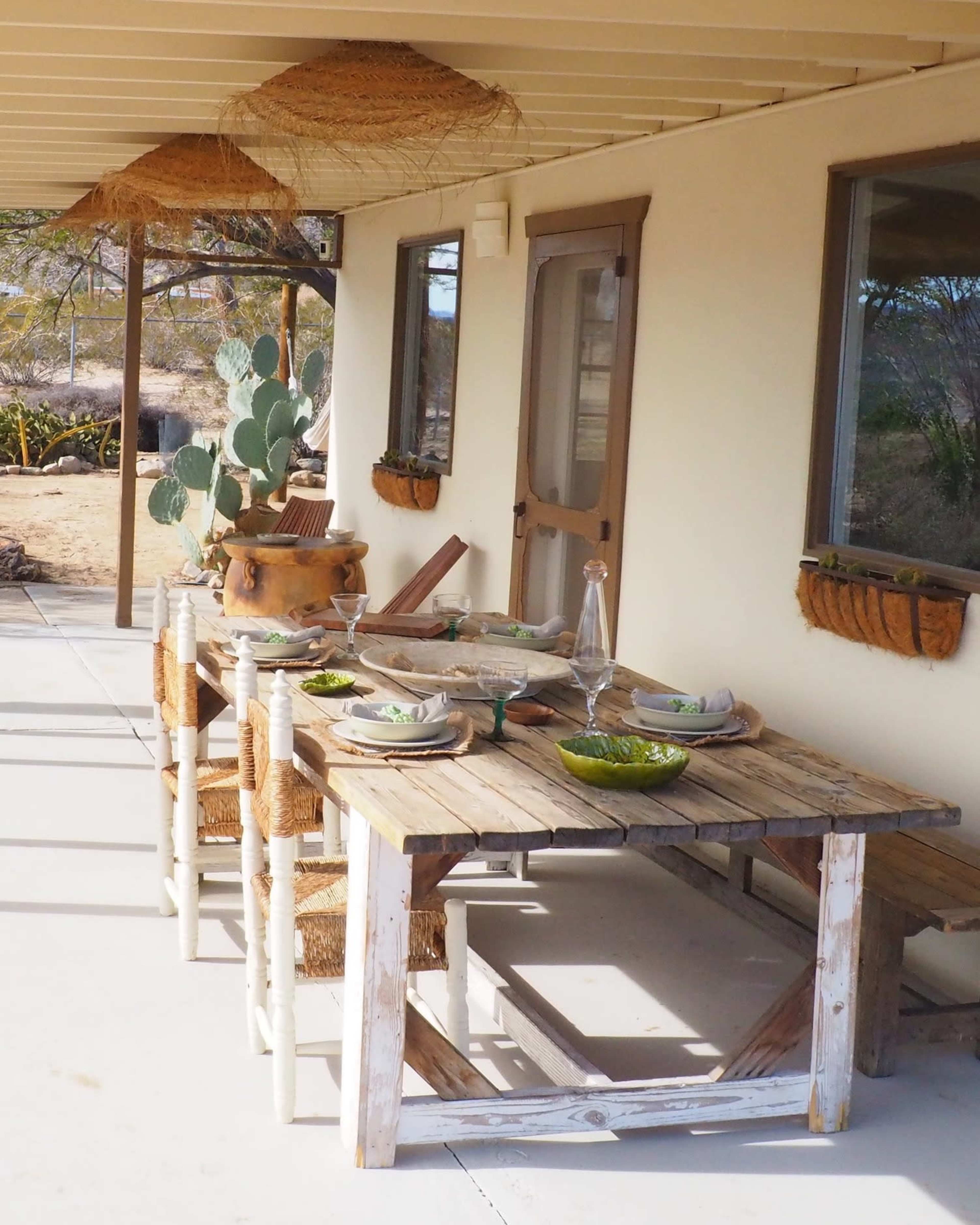A rustic wooden table set for a meal is positioned on a porch, surrounded by cactus plants and decorated with woven baskets.