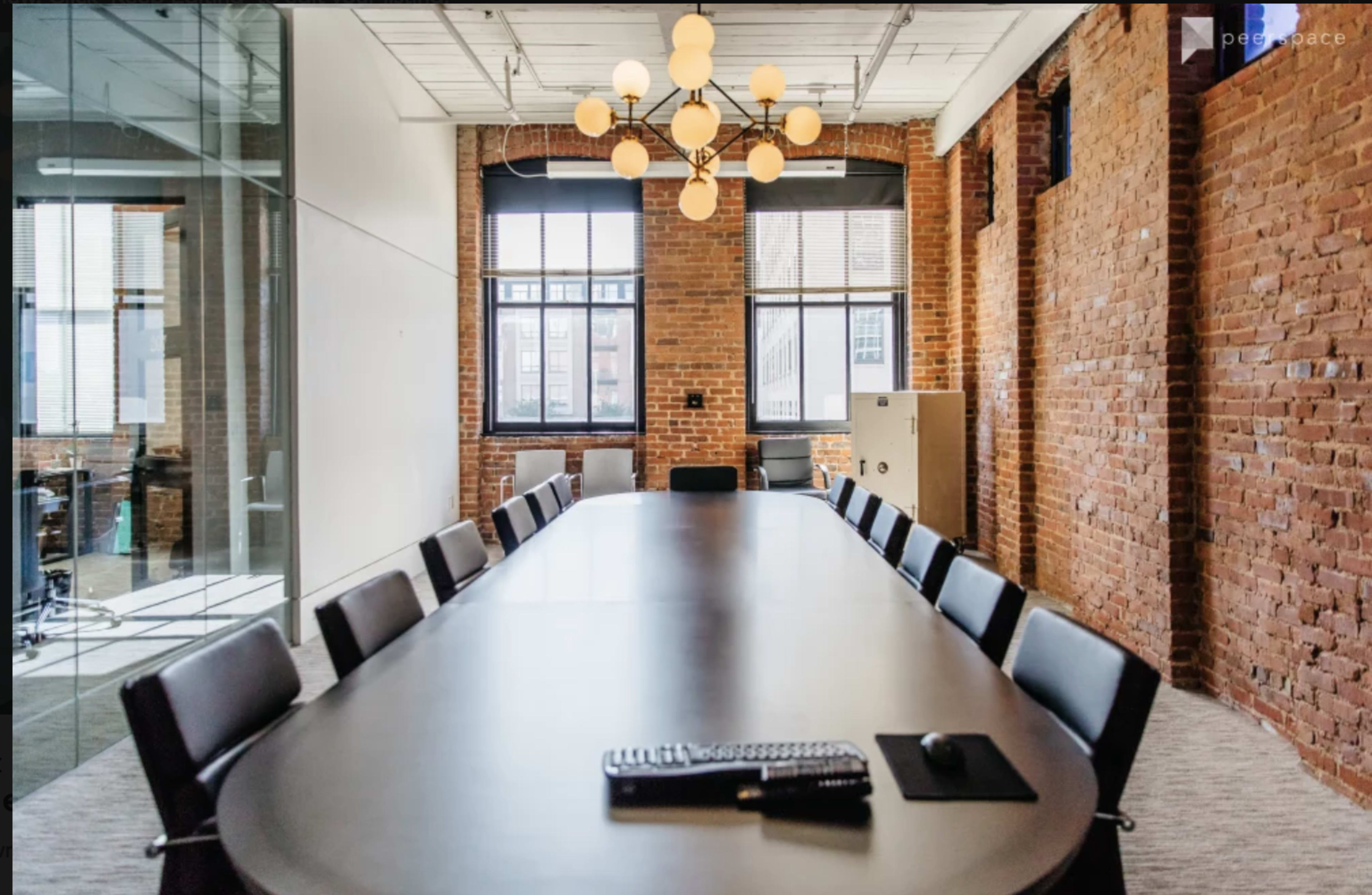 A long conference table with black chairs is set in a room featuring exposed brick walls and large windows, adorned with a chandelier overhead.