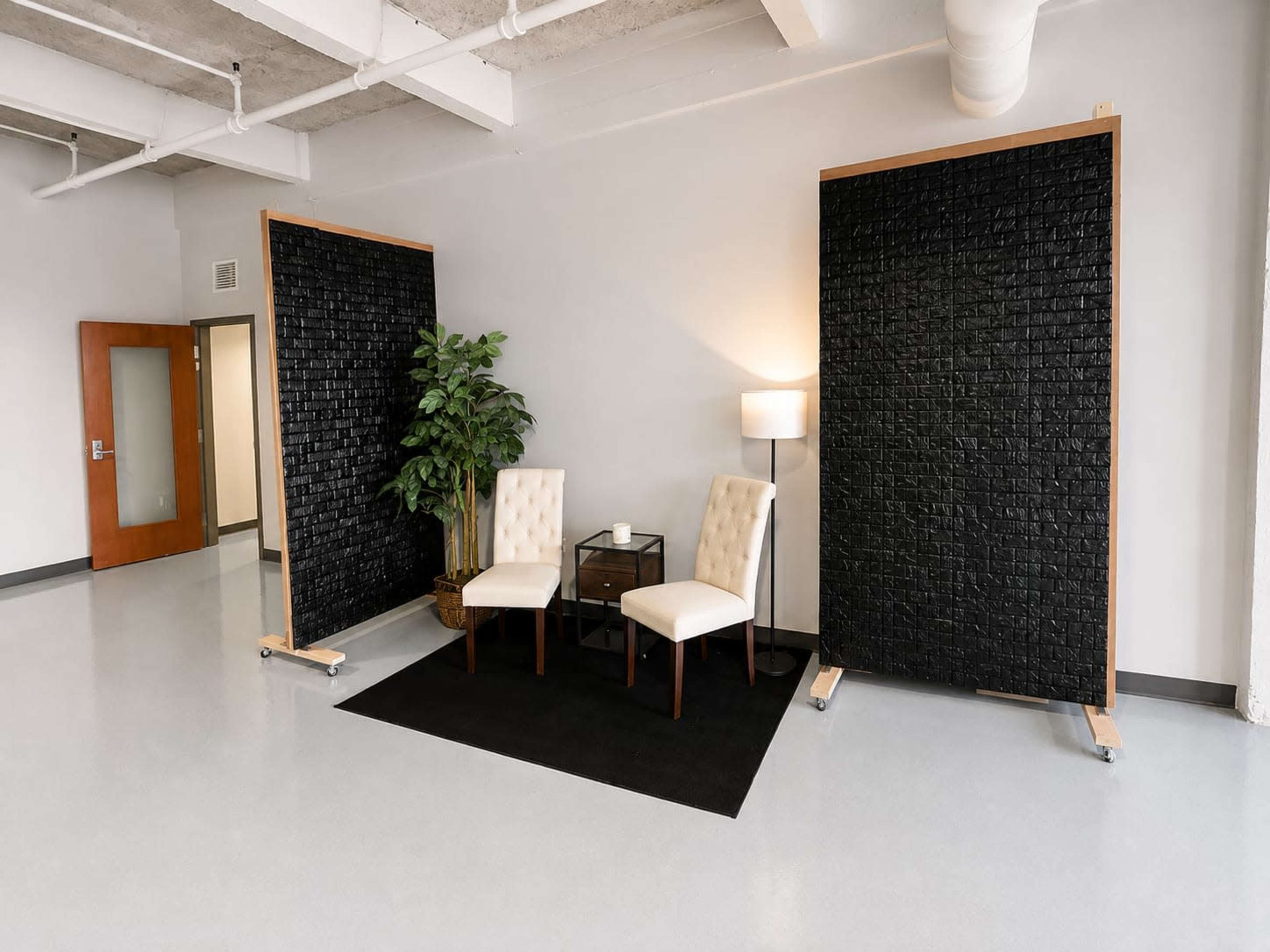 The image shows a minimalist seating area with two chairs, a small side table, a lamp, and a decorative plant, framed by two large black partitions.