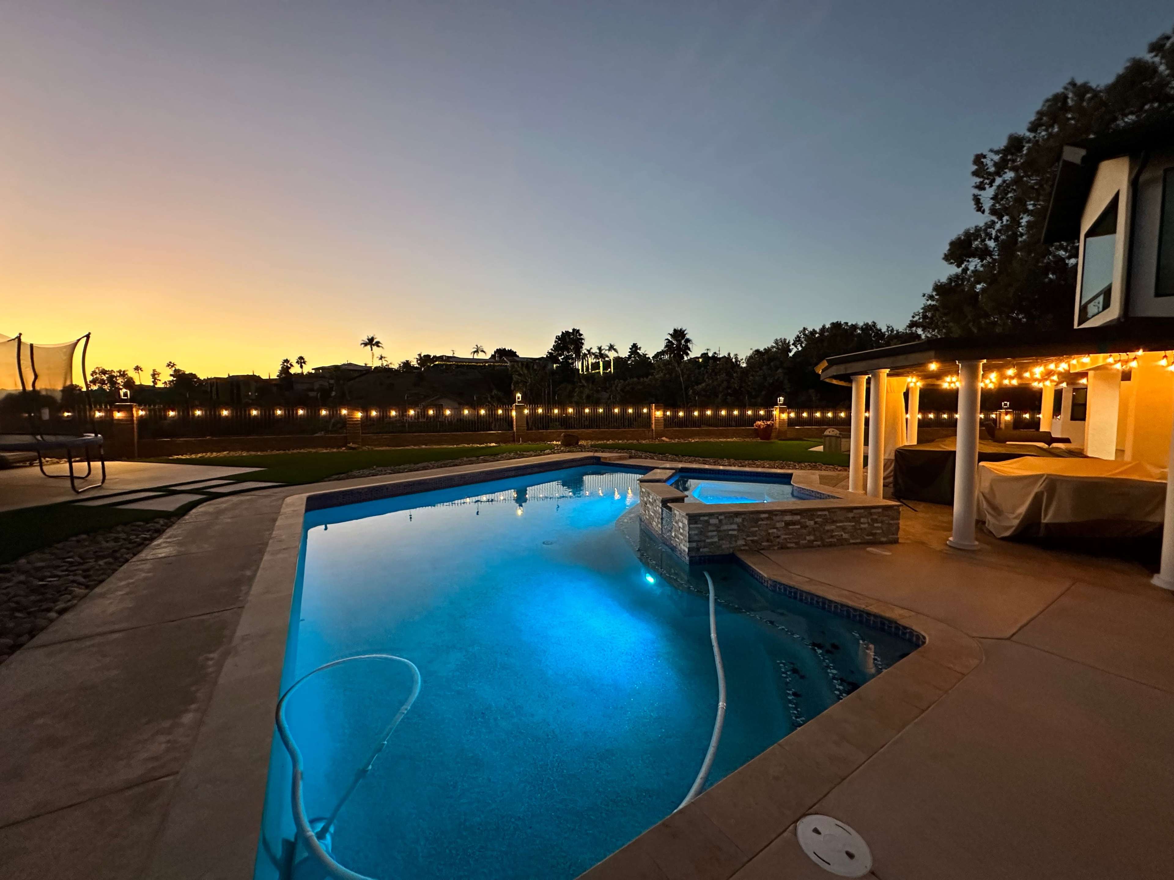A well-lit pool area features a blue swimming pool and spa surrounded by landscaped greenery at dusk.