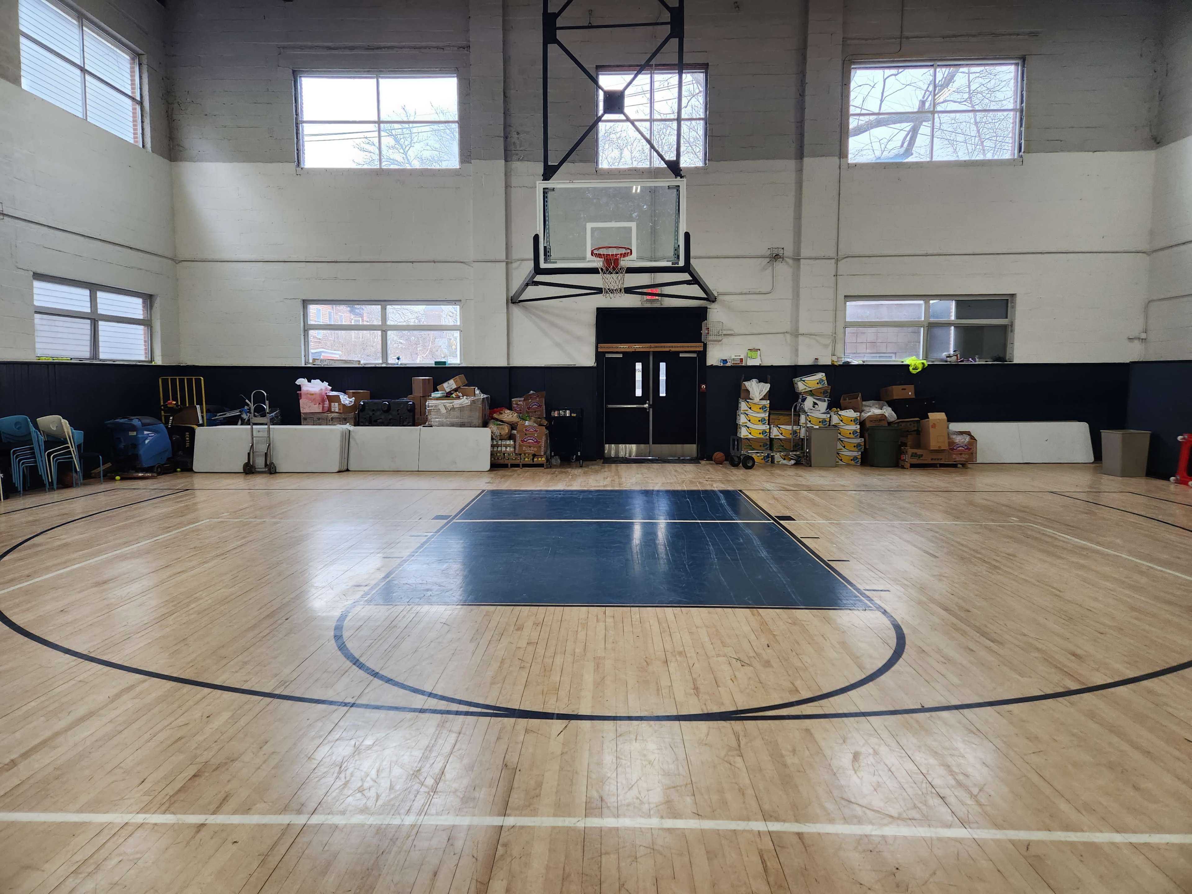 The image shows a basketball court with a hoop at one end and various items stacked against the walls.