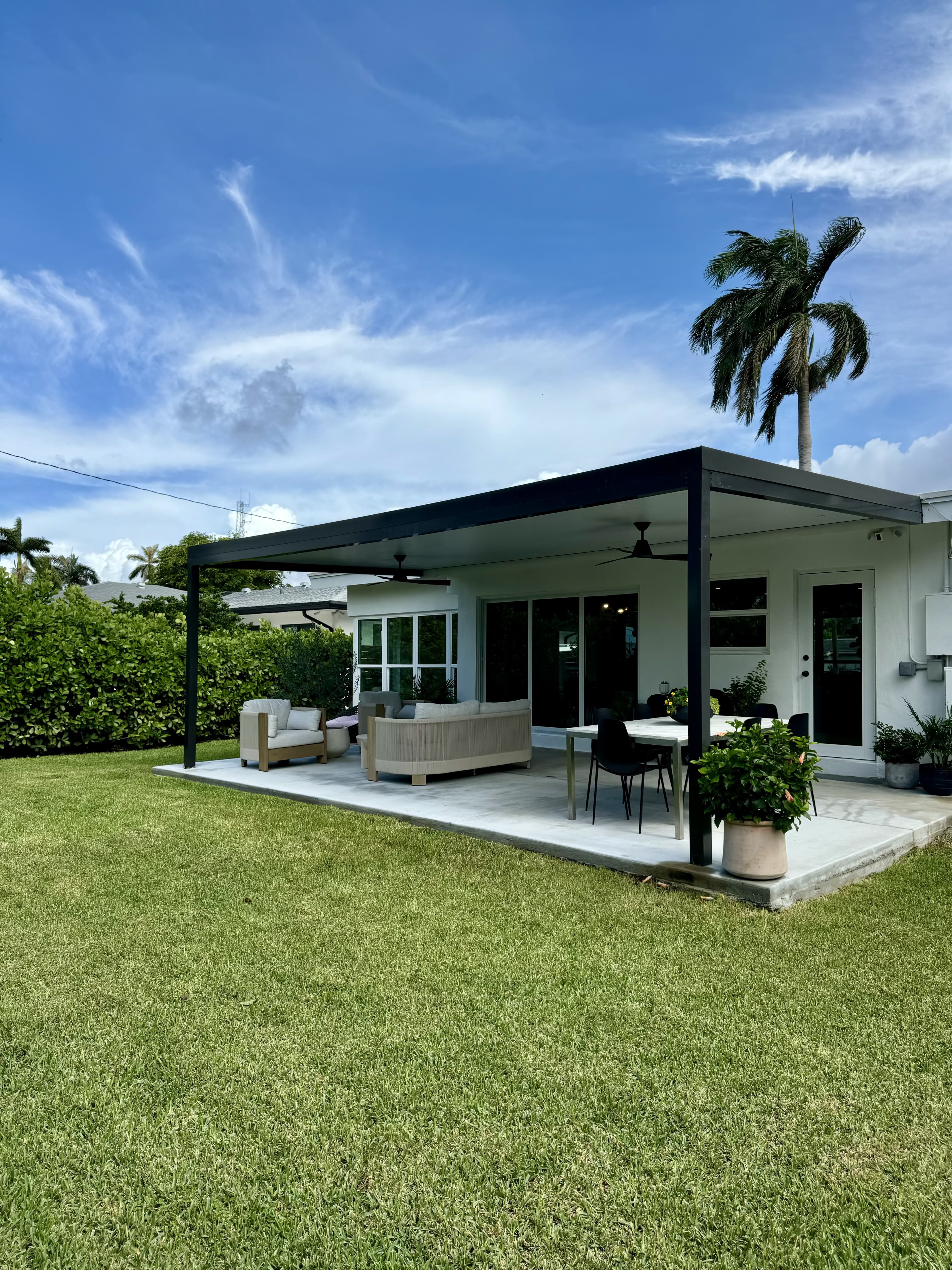 A modern patio with outdoor seating is situated behind a house, framed by lush greenery and palm trees under a clear blue sky.