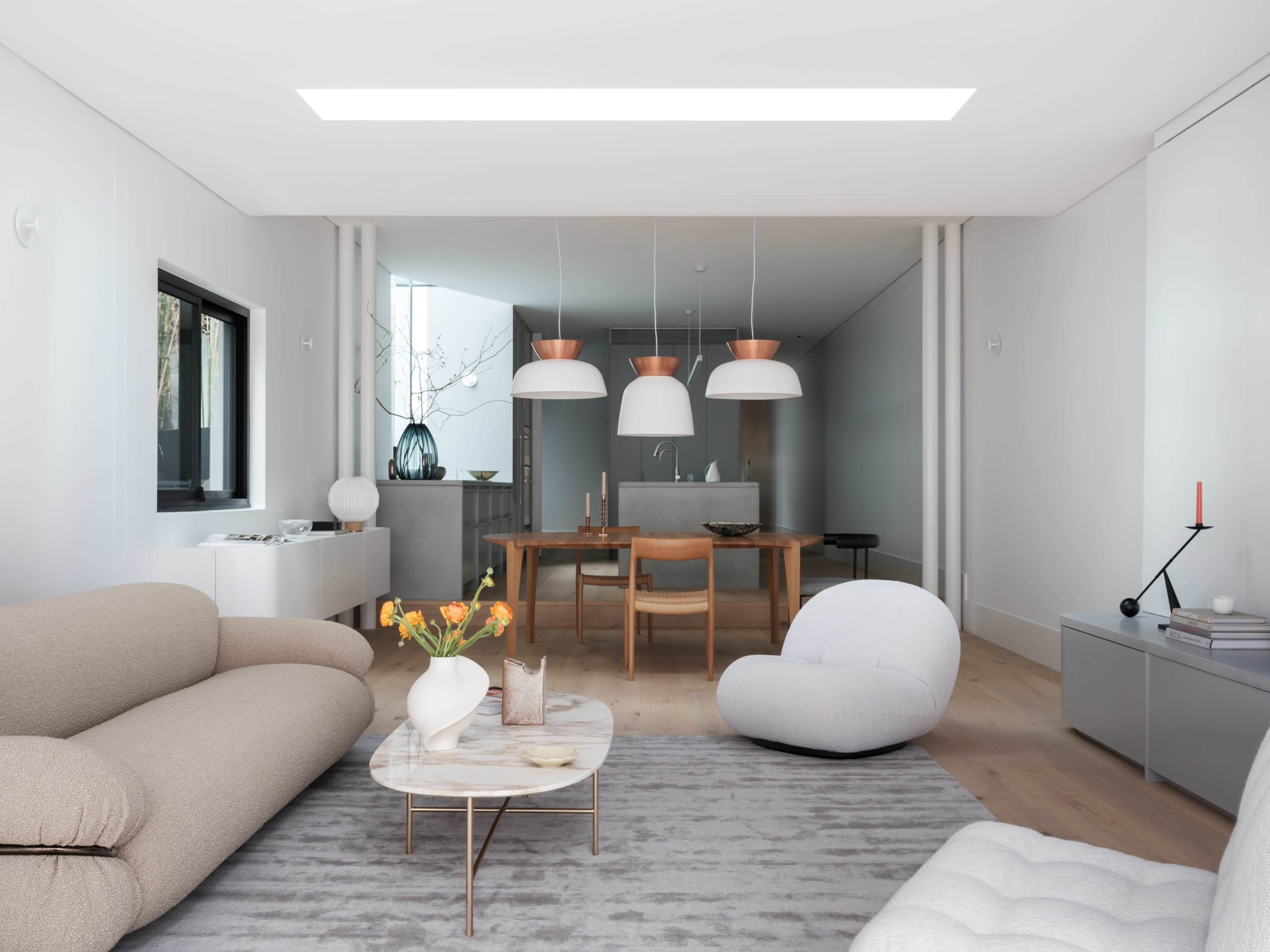A modern living room features a light-colored sofa, a round coffee table with a floral arrangement, and three pendant lights above a dining area with a large wooden table.