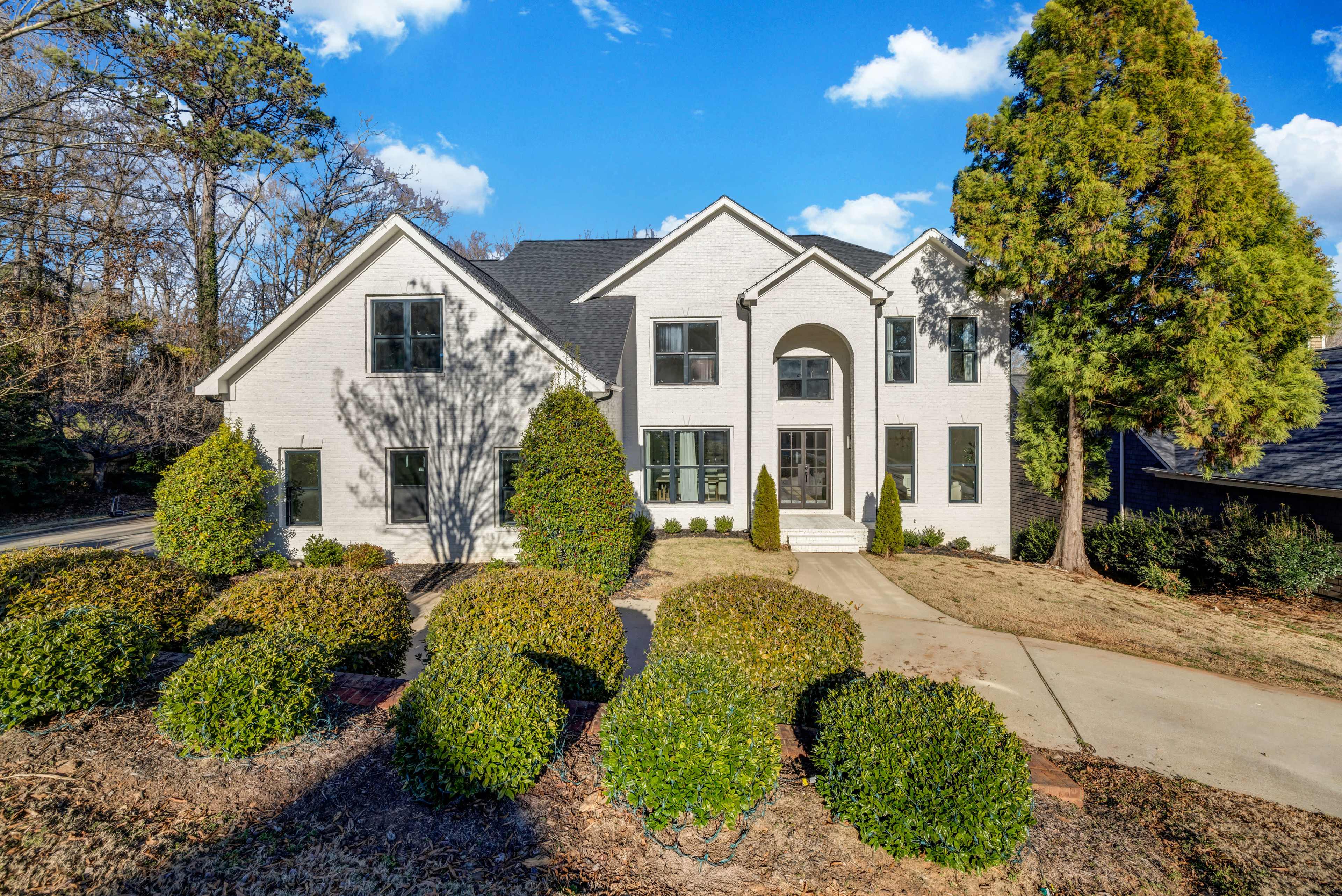 A large white brick house with multiple gables is surrounded by neatly trimmed hedges and a paved driveway under a blue sky.