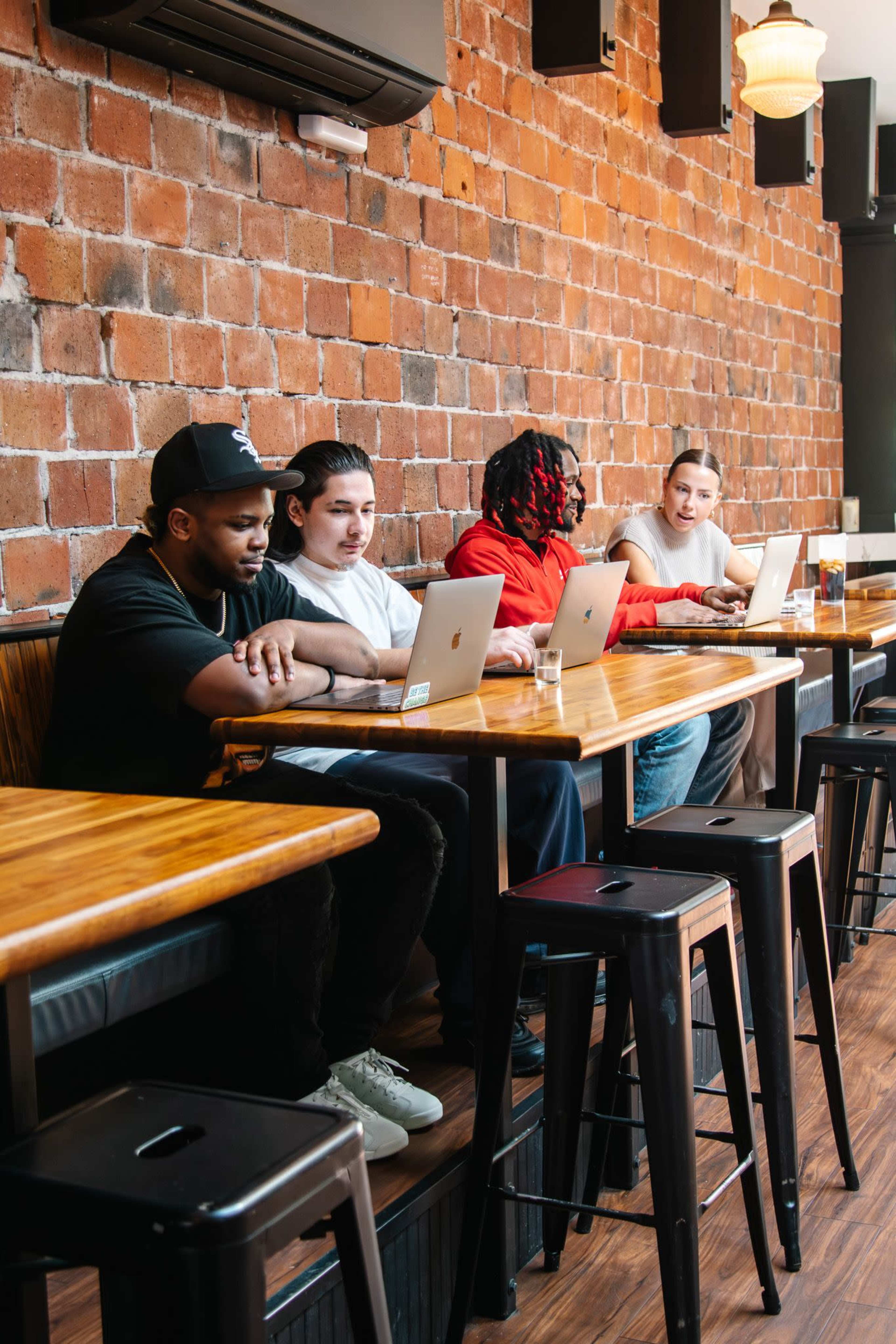 Four people are seated at a wooden bar in a cafe, each using laptops, with a brick wall in the background.