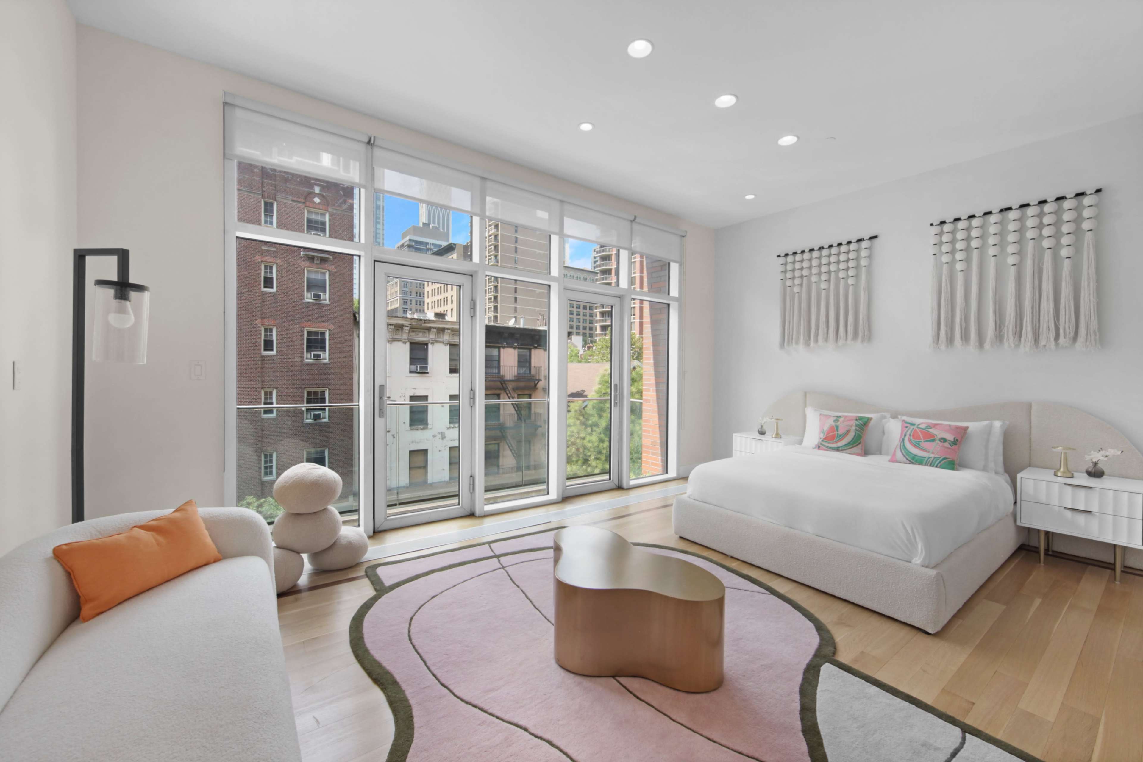 The image shows a modern bedroom with large windows, a bed, a sofa, and a round coffee table on a pink area rug.