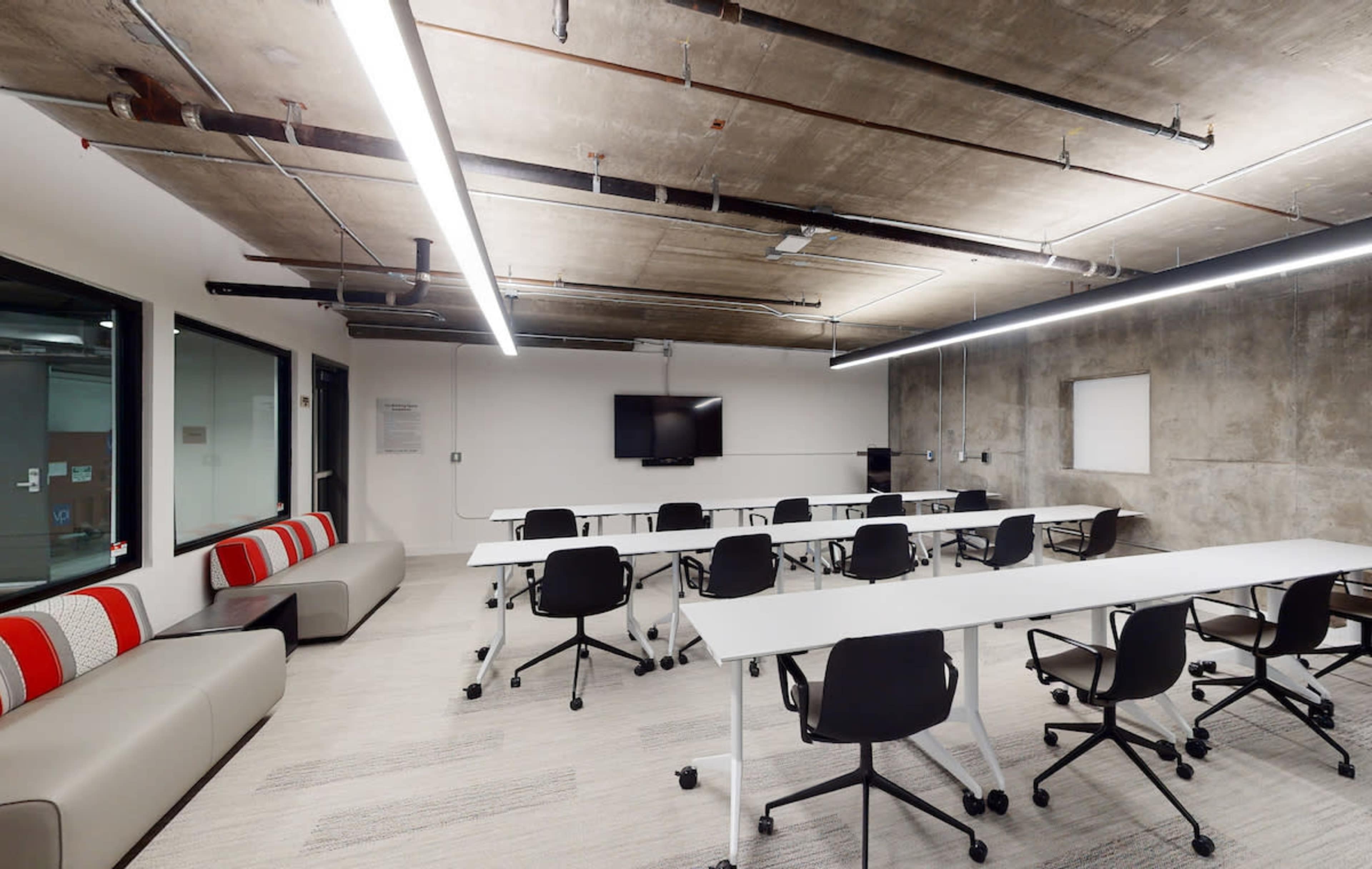 A modern conference room with rectangular tables arranged in a U-shape, black chairs, and large windows overlooking a concrete wall.