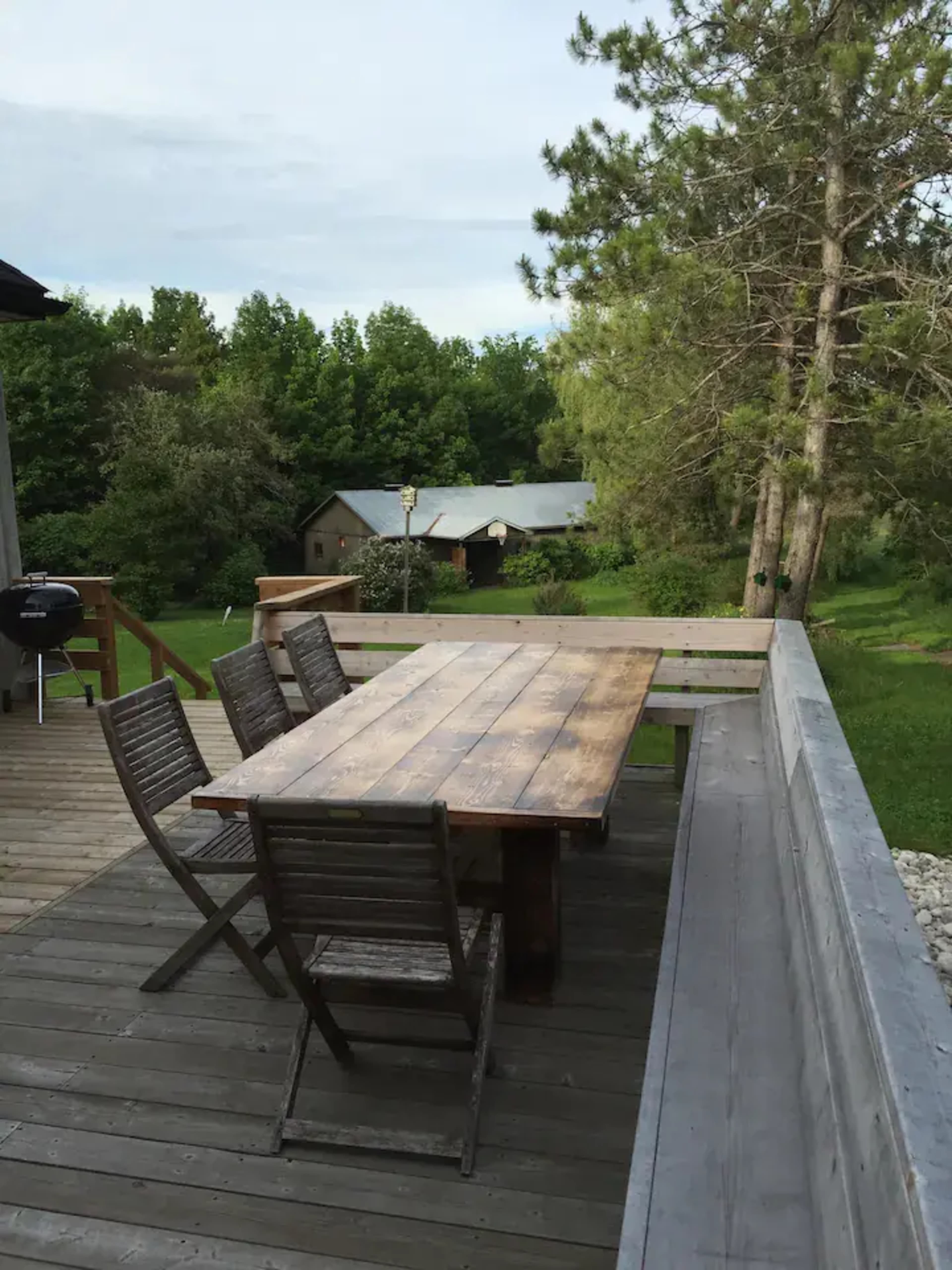 A wooden dining table and chairs are set on a deck overlooking a green landscape with trees and a distant building.