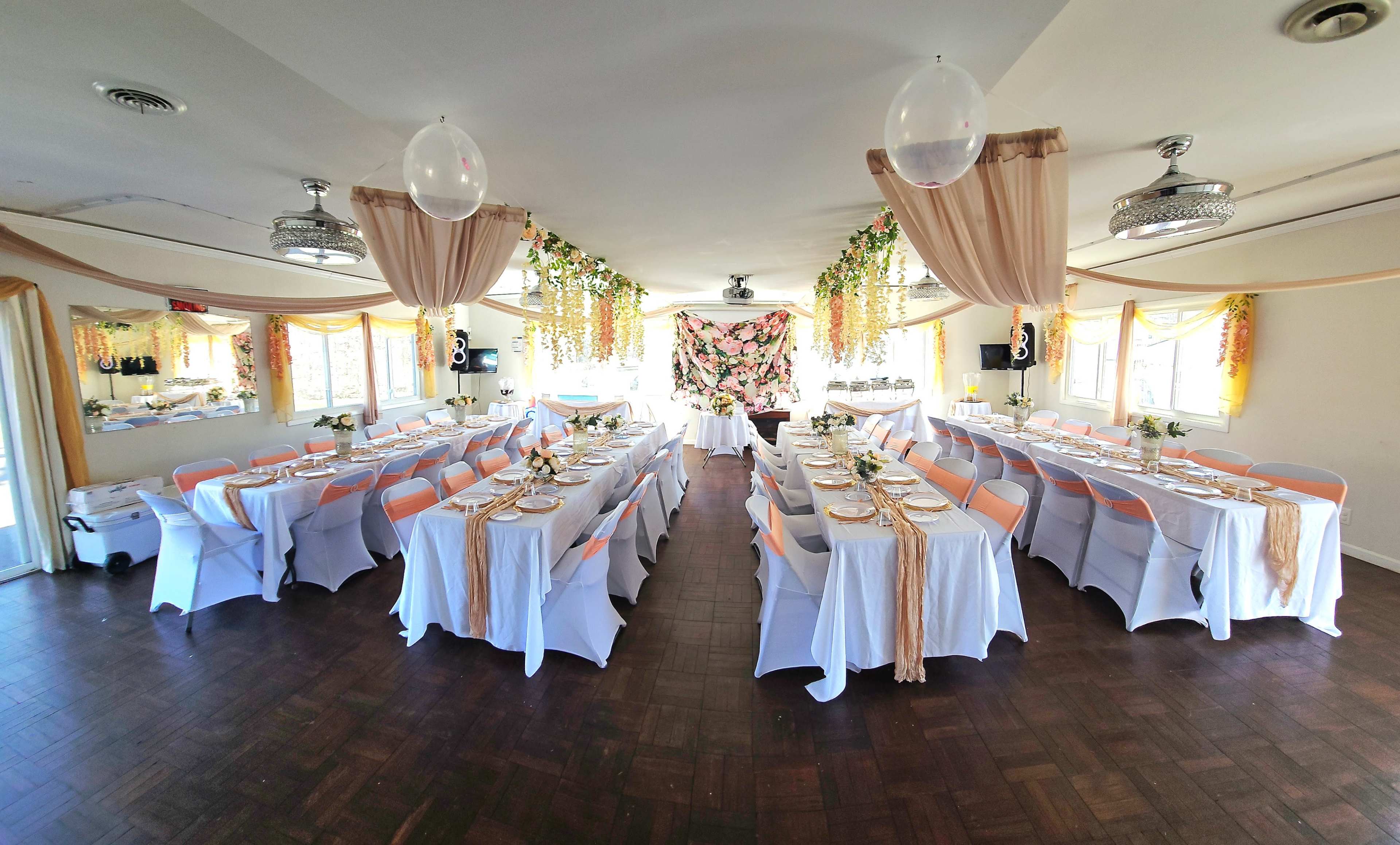 The image shows a banquet hall set up for a special event, featuring long tables covered with white tablecloths and decorated with floral arrangements, with additional drapery and a backdrop of flowers.