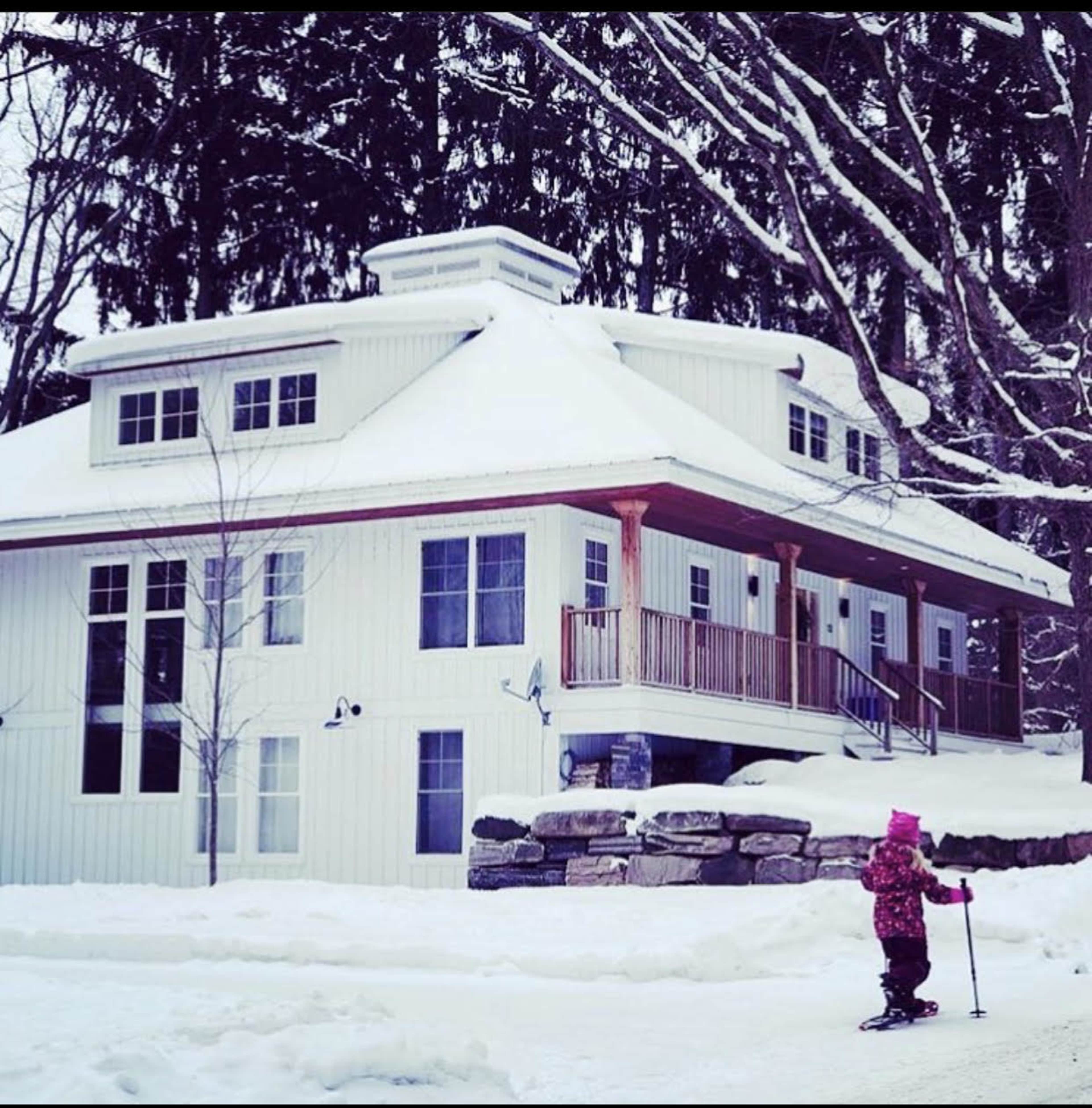A child in a pink hat skates on snow near a large white house with a wraparound porch and stone foundation.