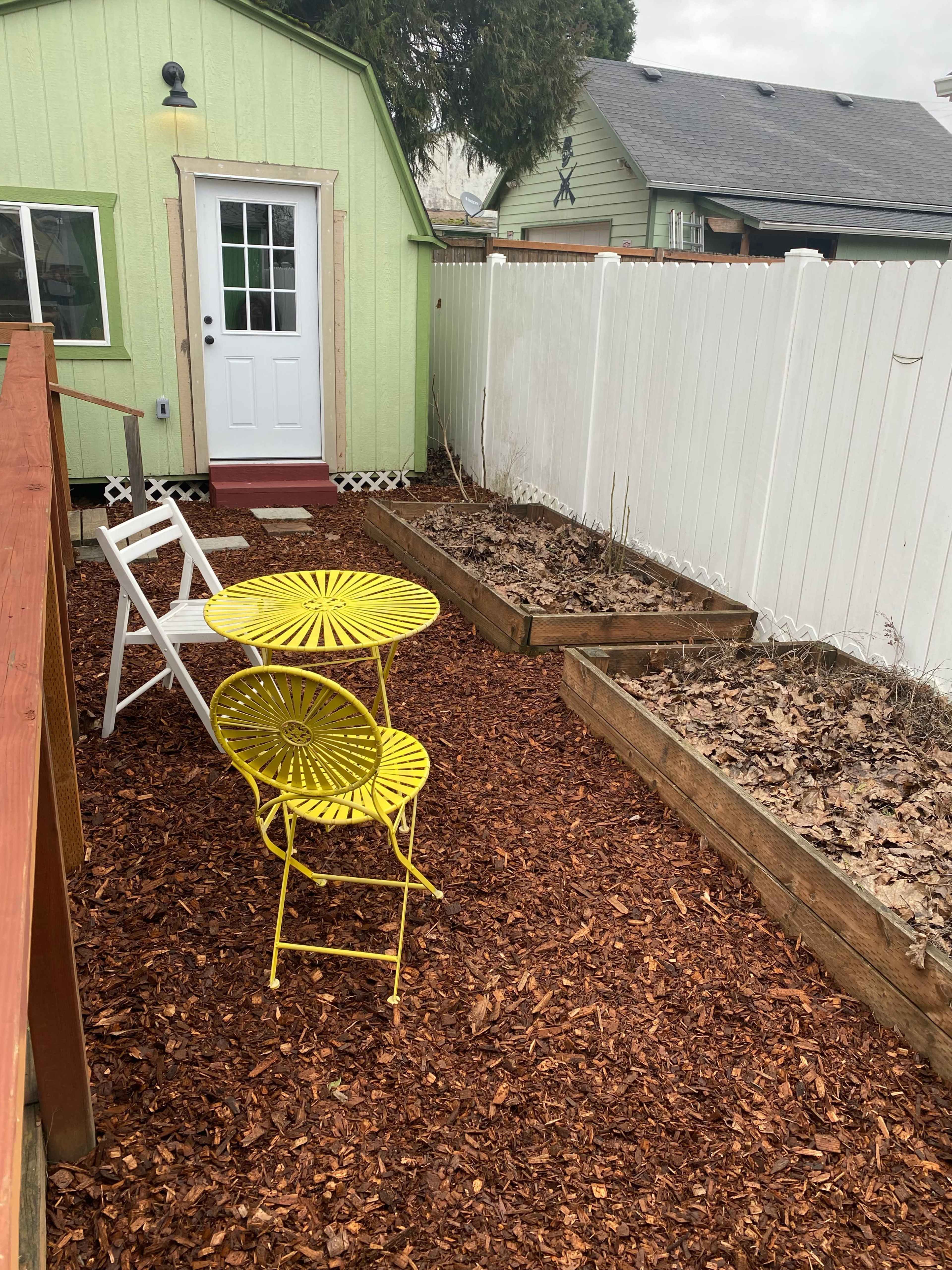 A small patio area features a yellow bistro table and chairs surrounded by wooden garden beds and a white fence.