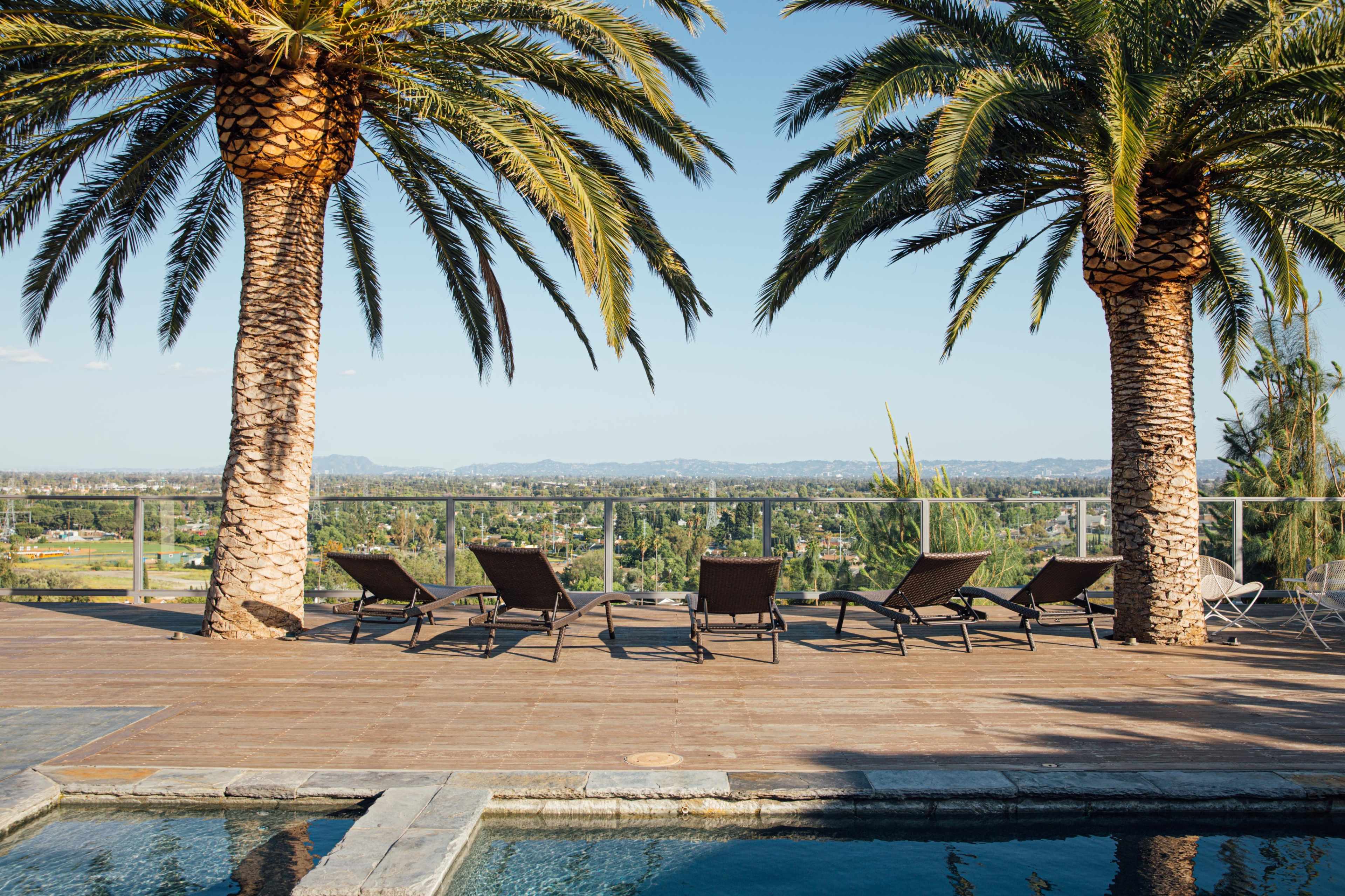 A poolside area with lounge chairs positioned under tall palm trees, overlooking a scenic landscape.