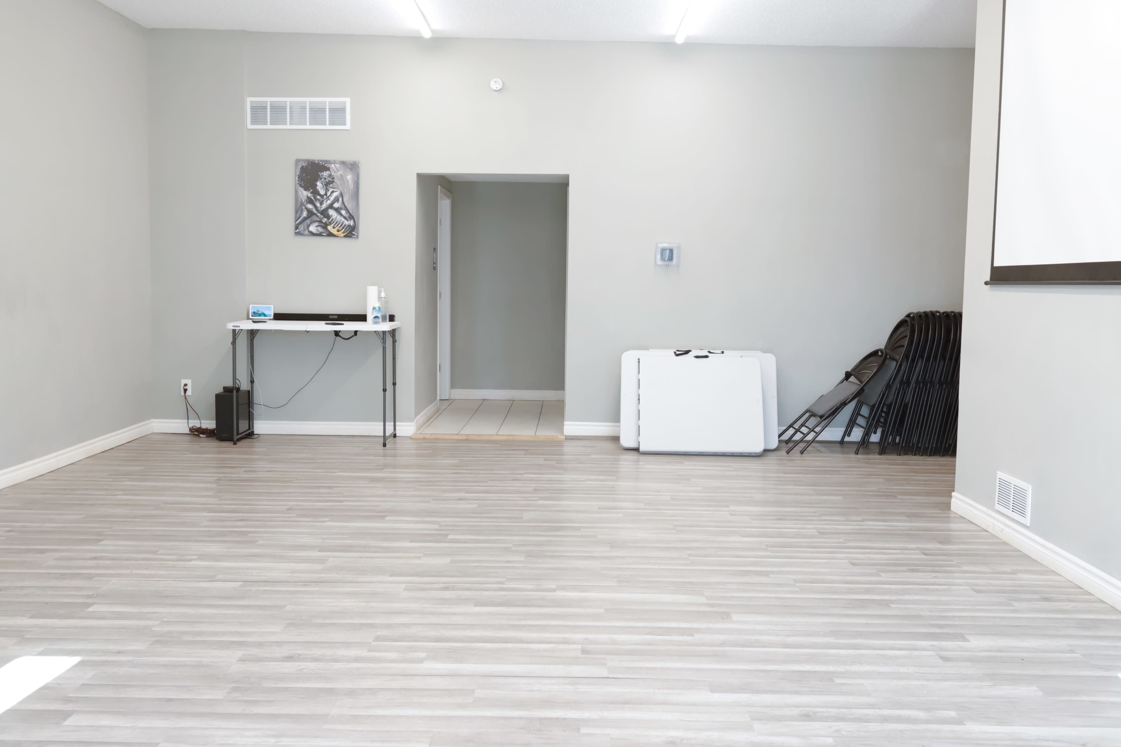 A spacious, well-lit room with light-colored wood flooring, a table with a few items on it, and a doorway leading to another area, along with some stacked chairs against the wall.