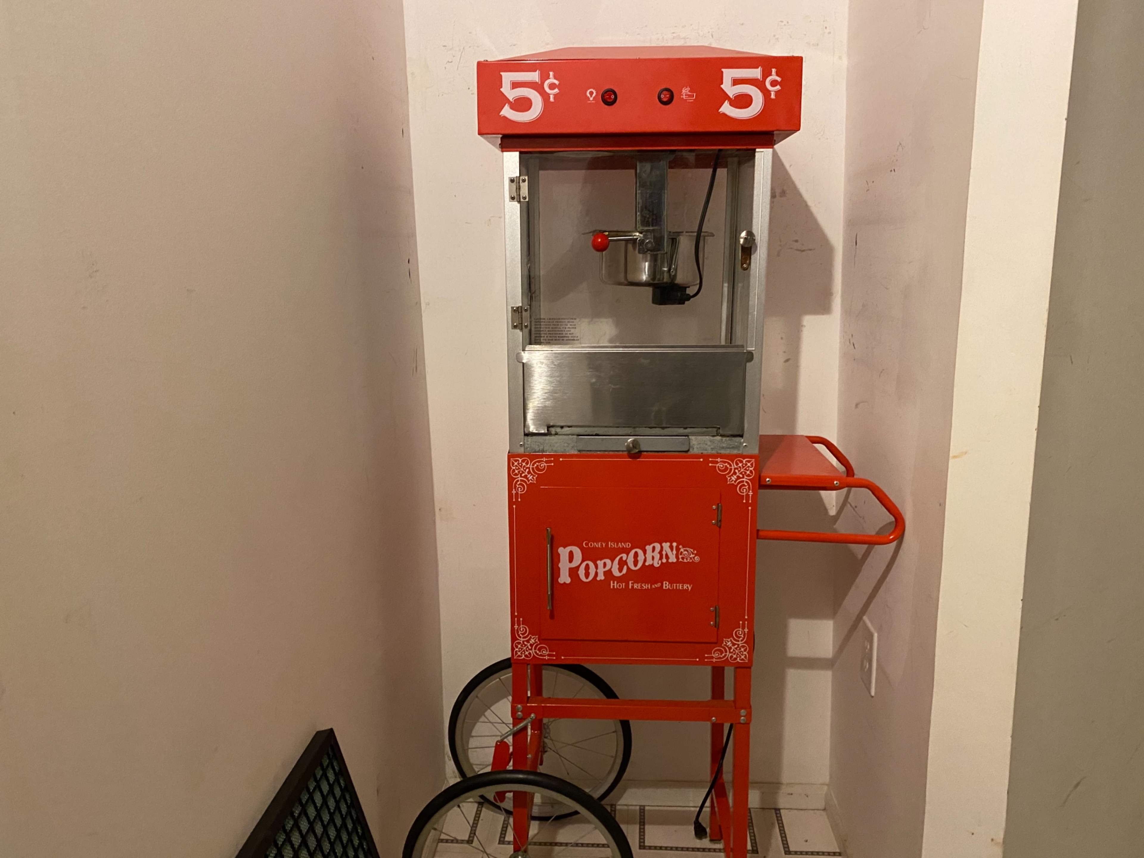 A vintage popcorn machine with a red exterior and a price of five cents displayed on the front is positioned against a wall.