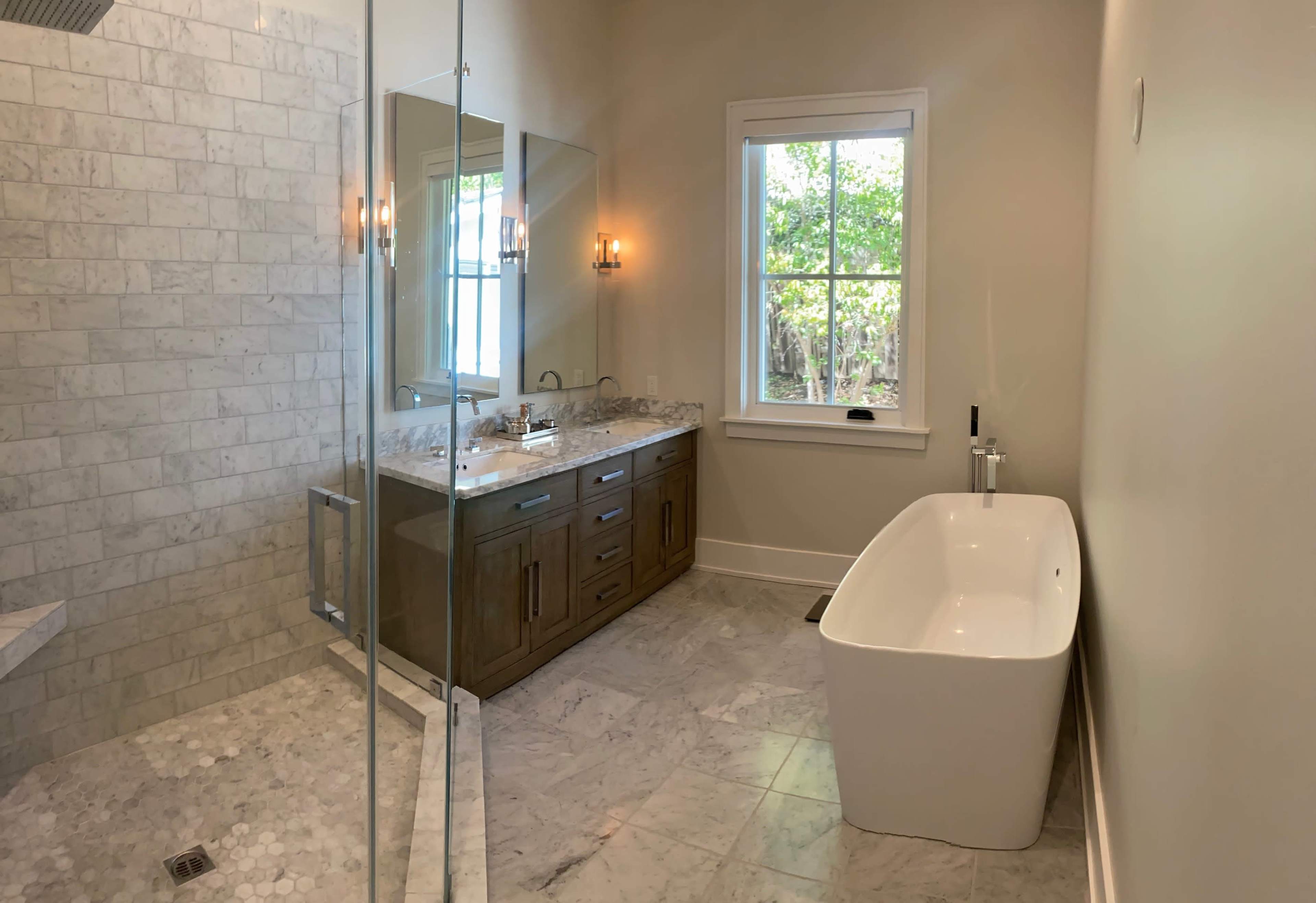 A modern bathroom featuring a glass-enclosed shower, a freestanding bathtub, a dual sink vanity with a granite countertop, and large windows providing natural light.
