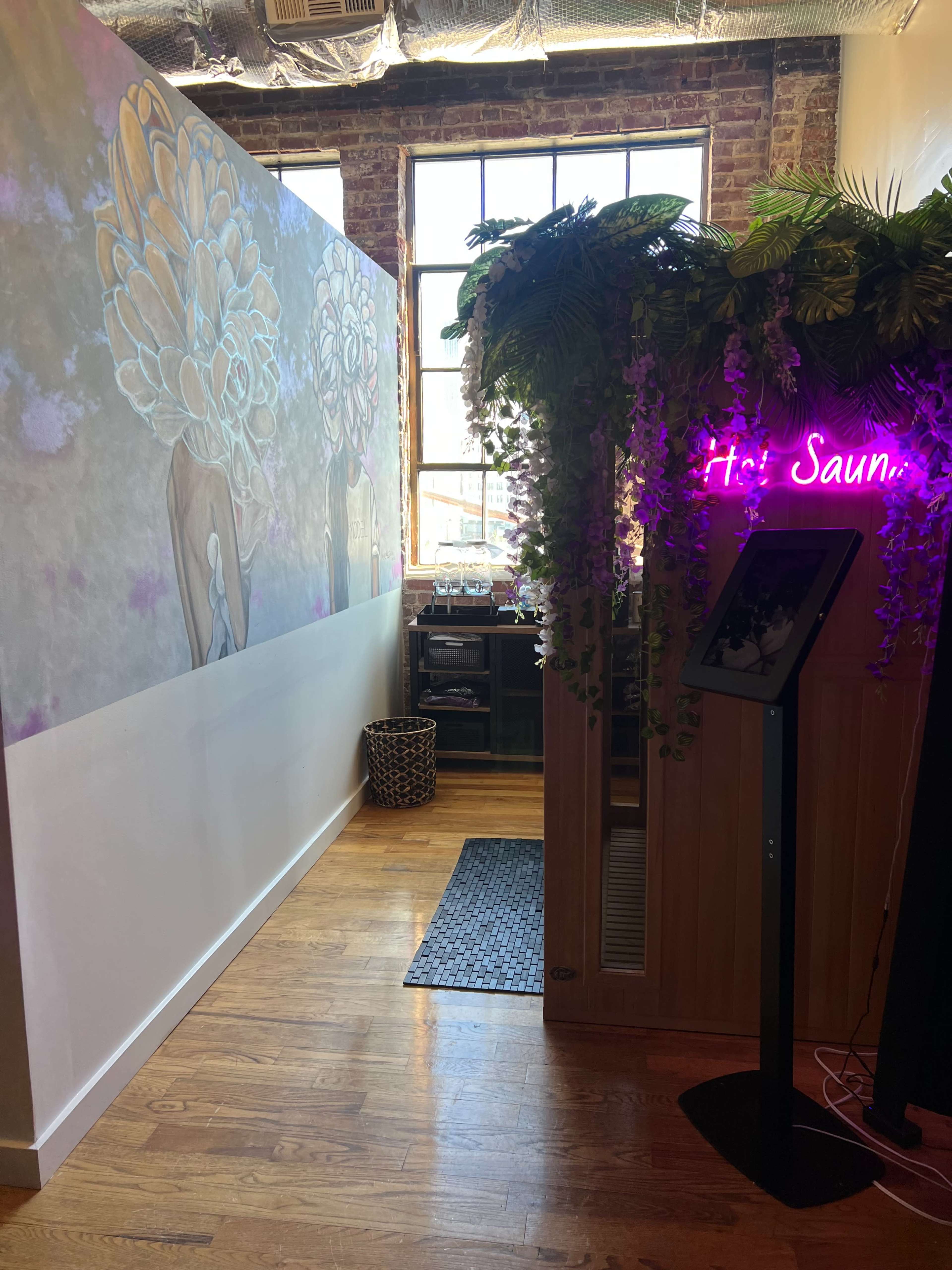 A bright hallway leading to a sauna, decorated with a mural and plants, and featuring a neon sign that reads "It's Sauna."