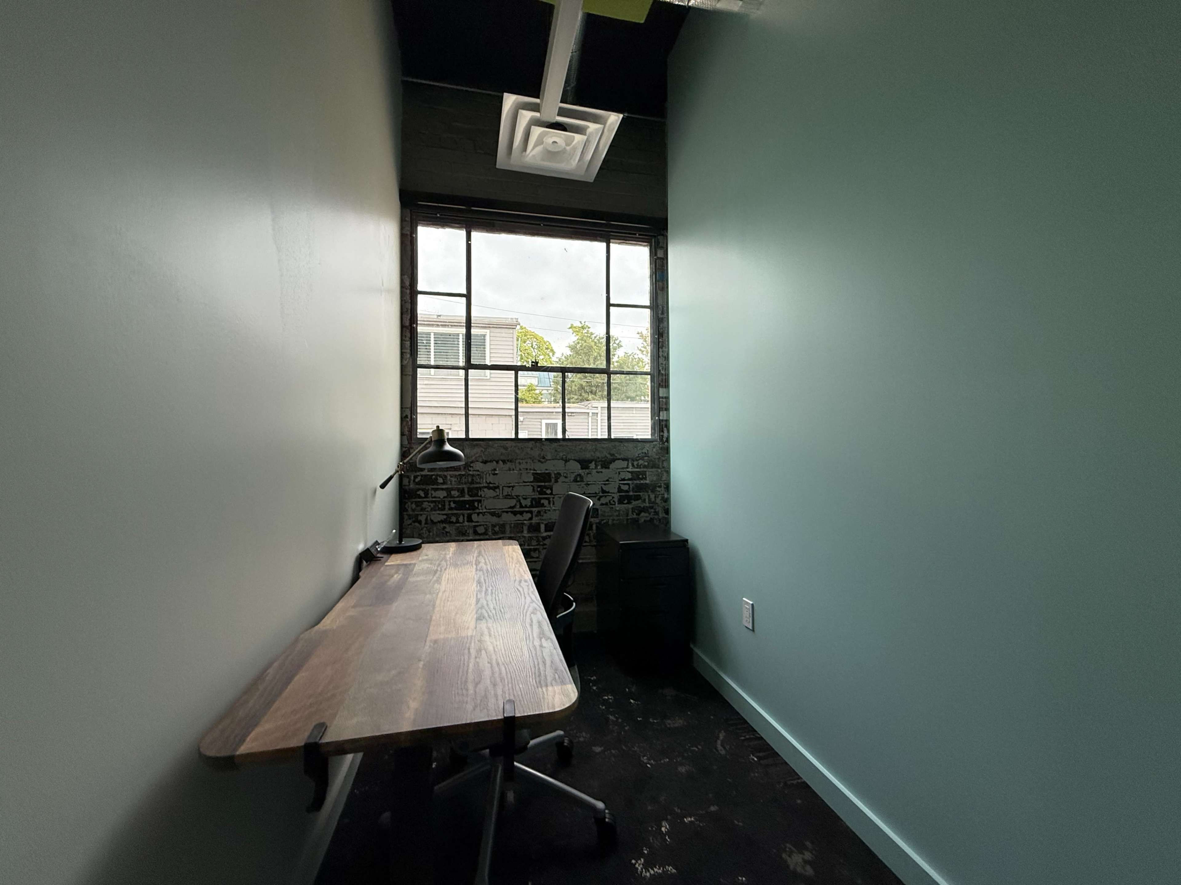 The image shows a narrow room with a wooden desk, an office chair, and a large window that partially illuminates the space.