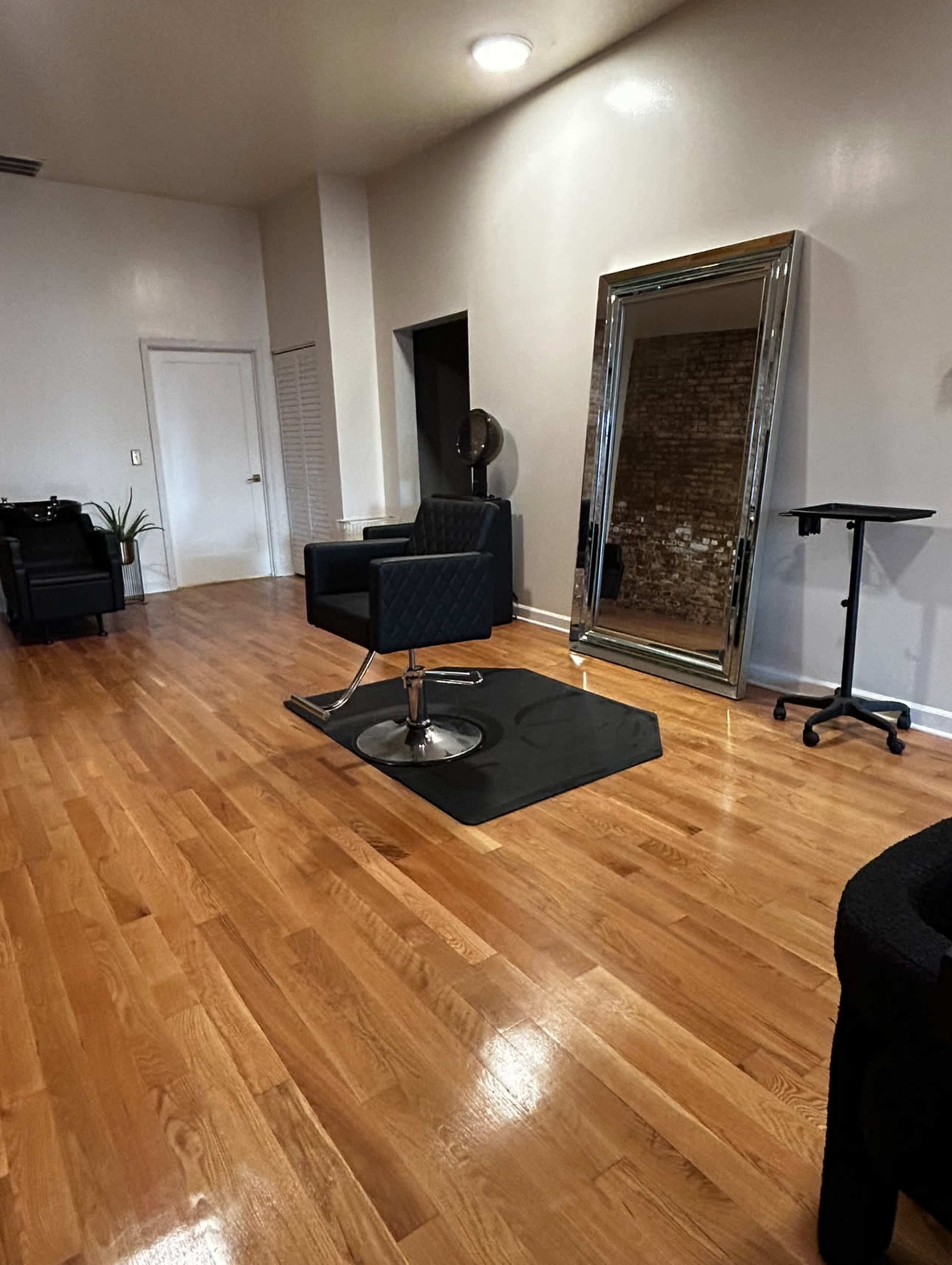A spacious salon interior with a styling chair on a black mat, a large mirror, and minimalist furniture against light-colored walls and hardwood flooring.