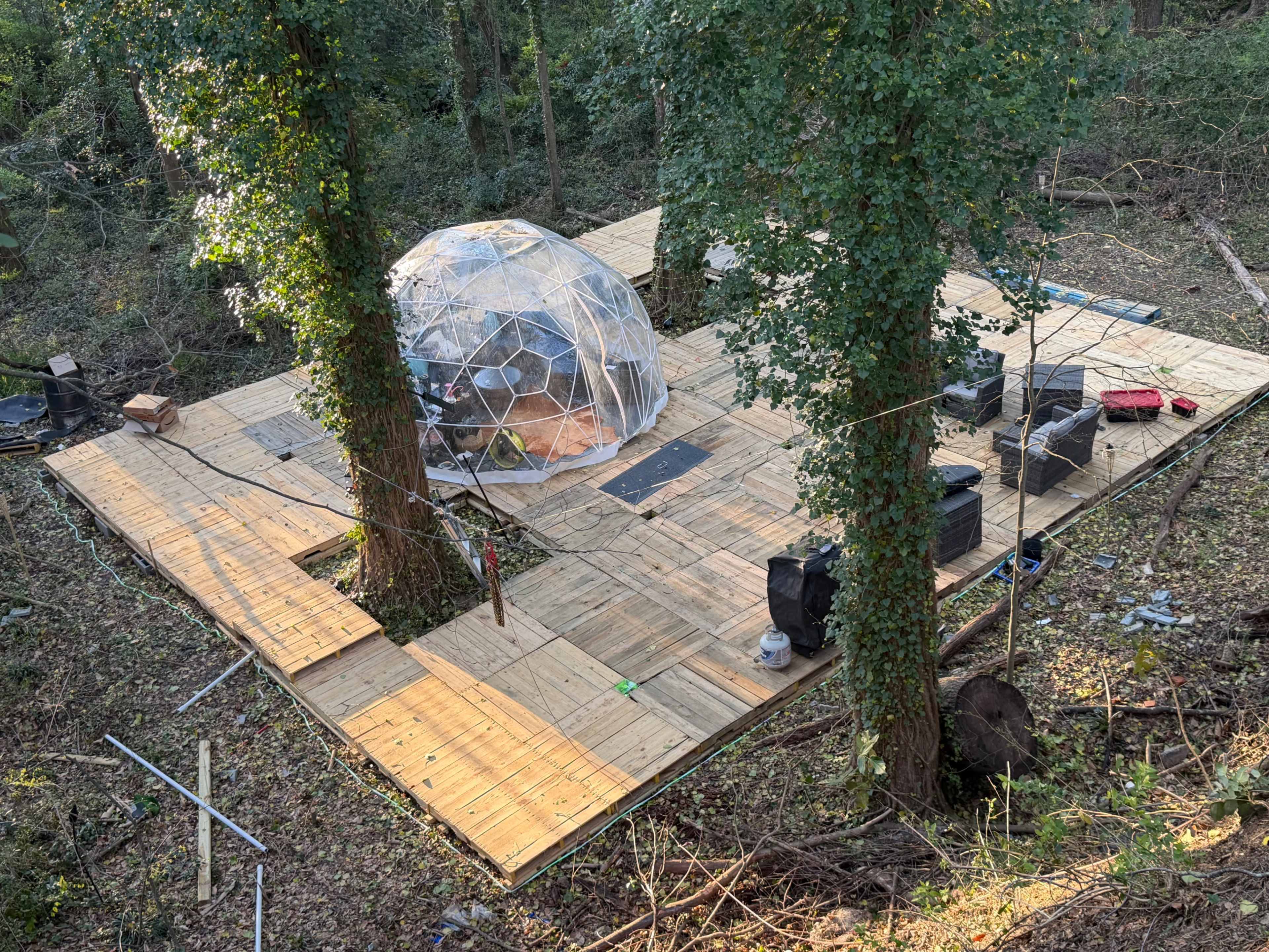 A geodesic dome sits on a wooden platform in a wooded area, surrounded by trees and construction materials.