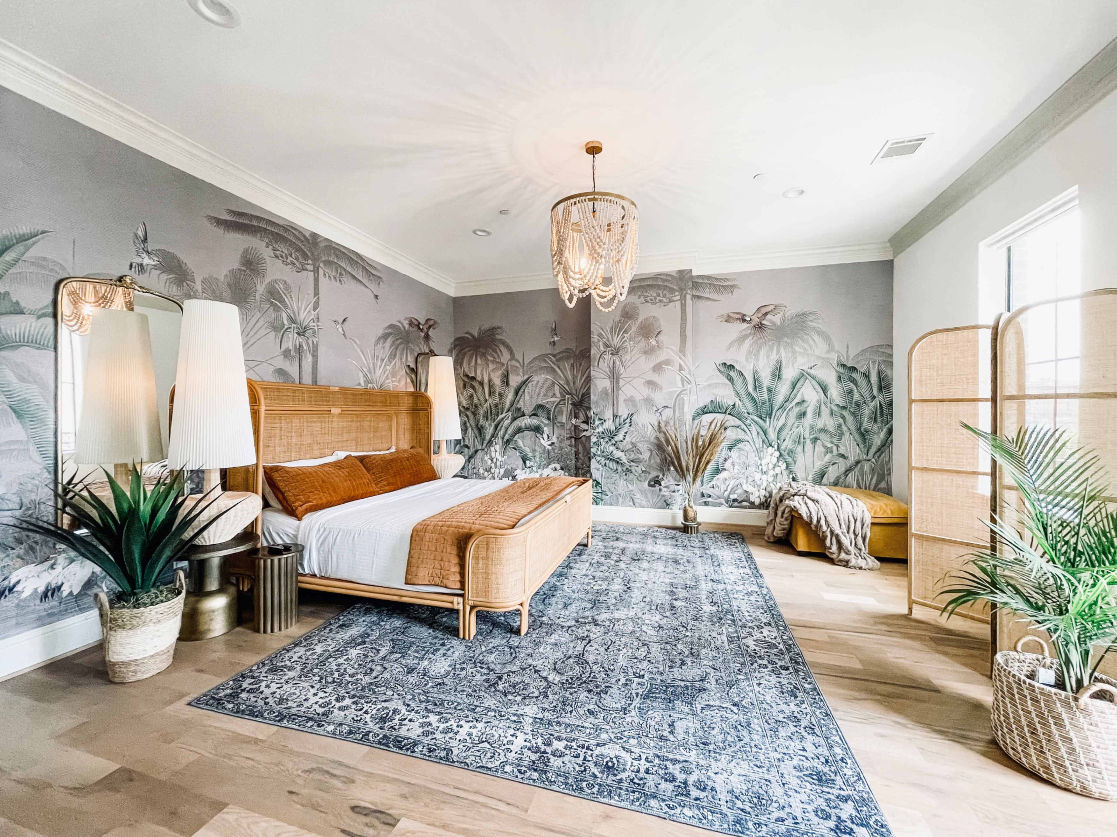 A spacious bedroom features a rattan bed, tropical mural walls, decorative plants, and a woven area rug.