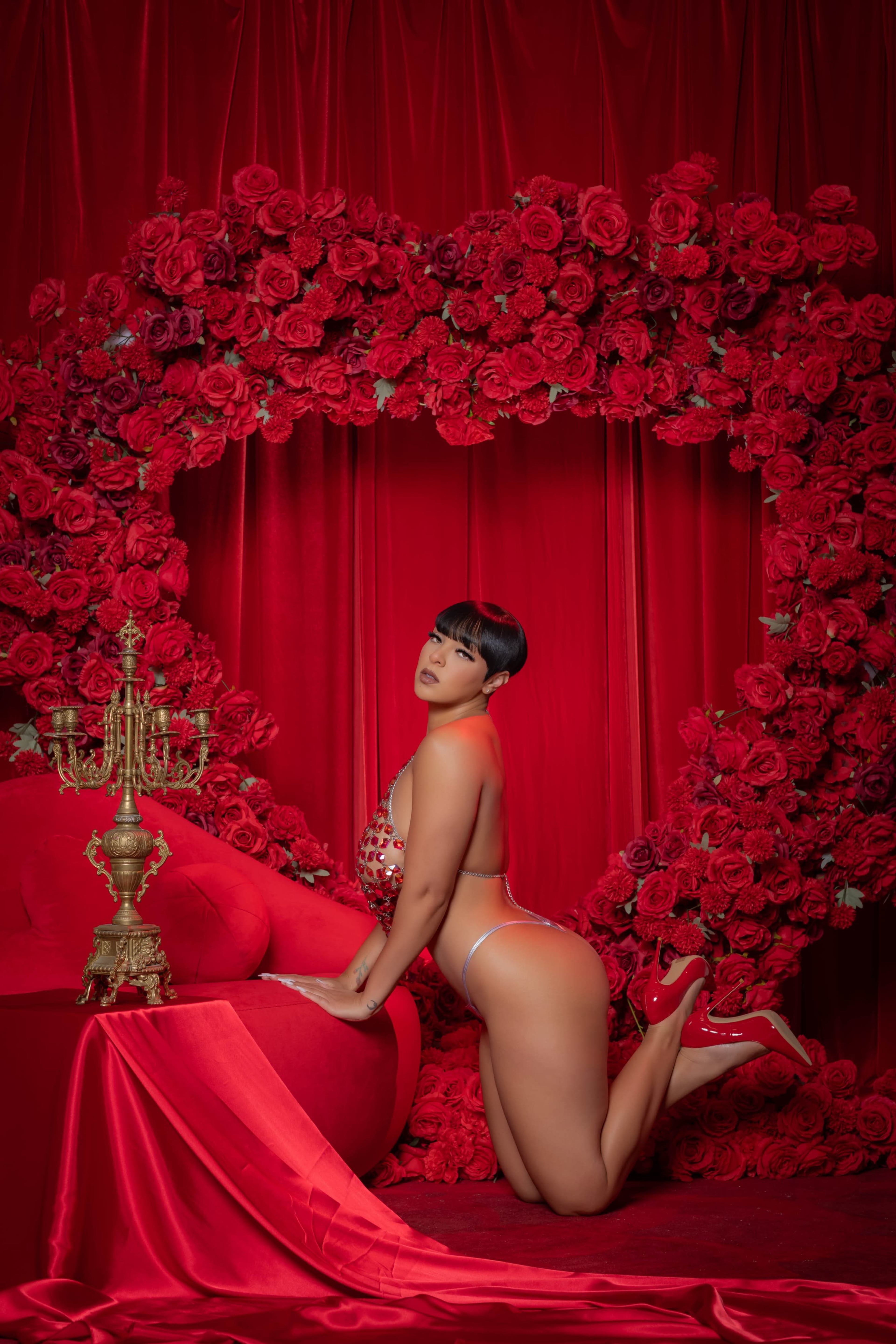 A person poses on their knees in front of a heart-shaped arrangement of red roses, with a candelabra on a red draped surface.