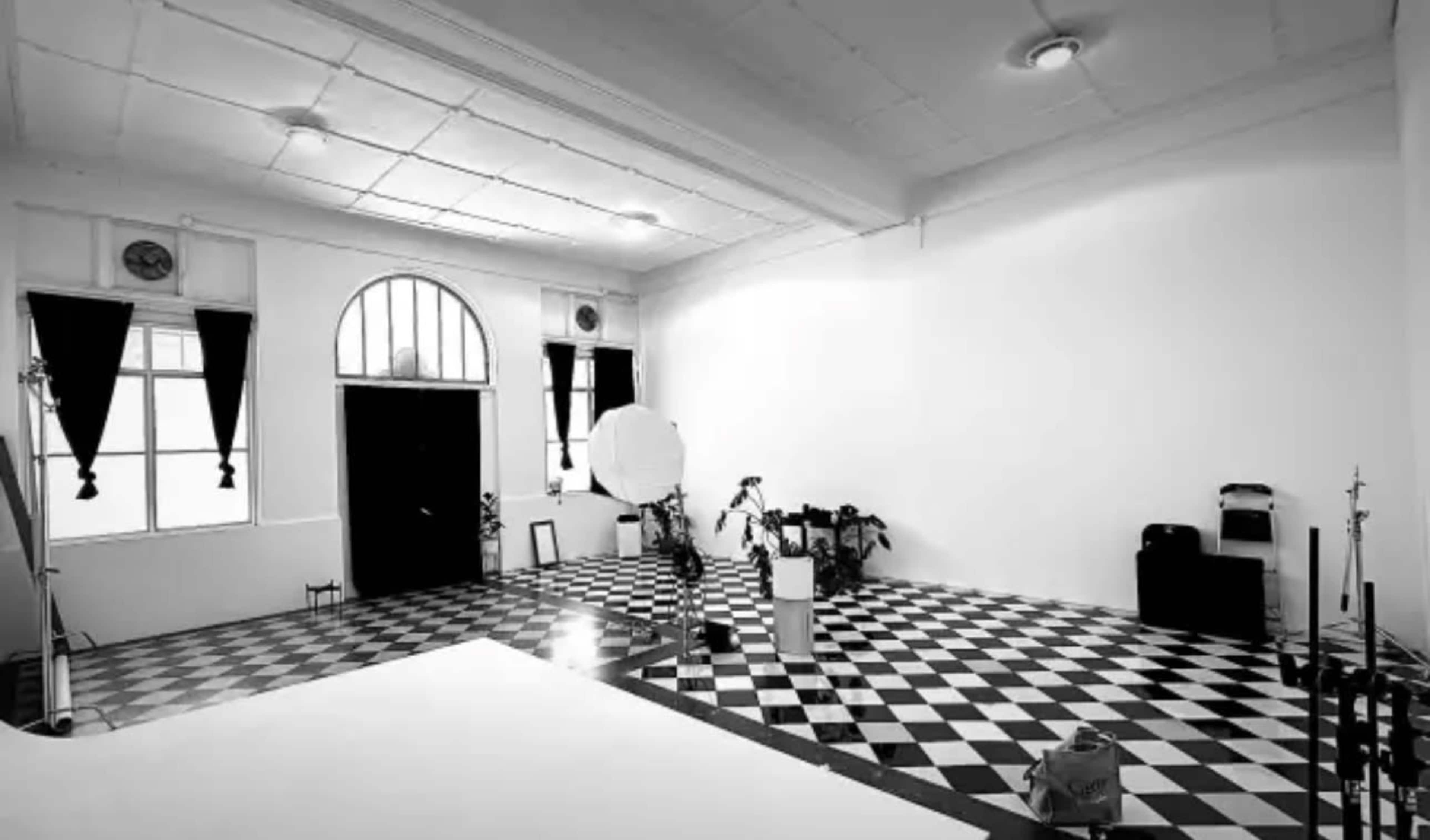 The image shows a spacious studio with black and white checkered flooring, large windows, and minimal furniture including a photography backdrop and equipment.
