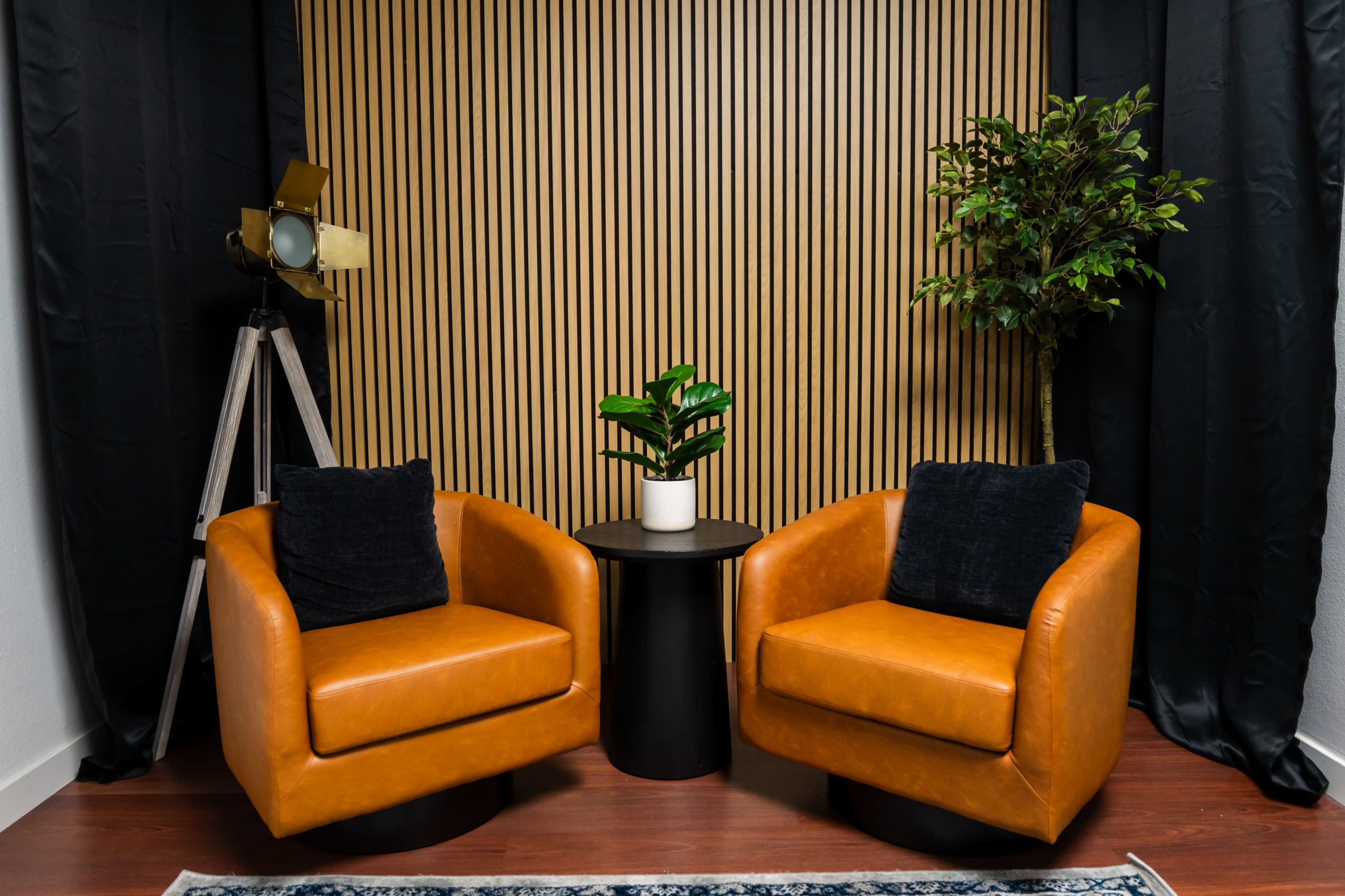 The image shows two leather chairs facing each other with a small black table between them, set against a backdrop of vertical wooden slats and dark drapes.