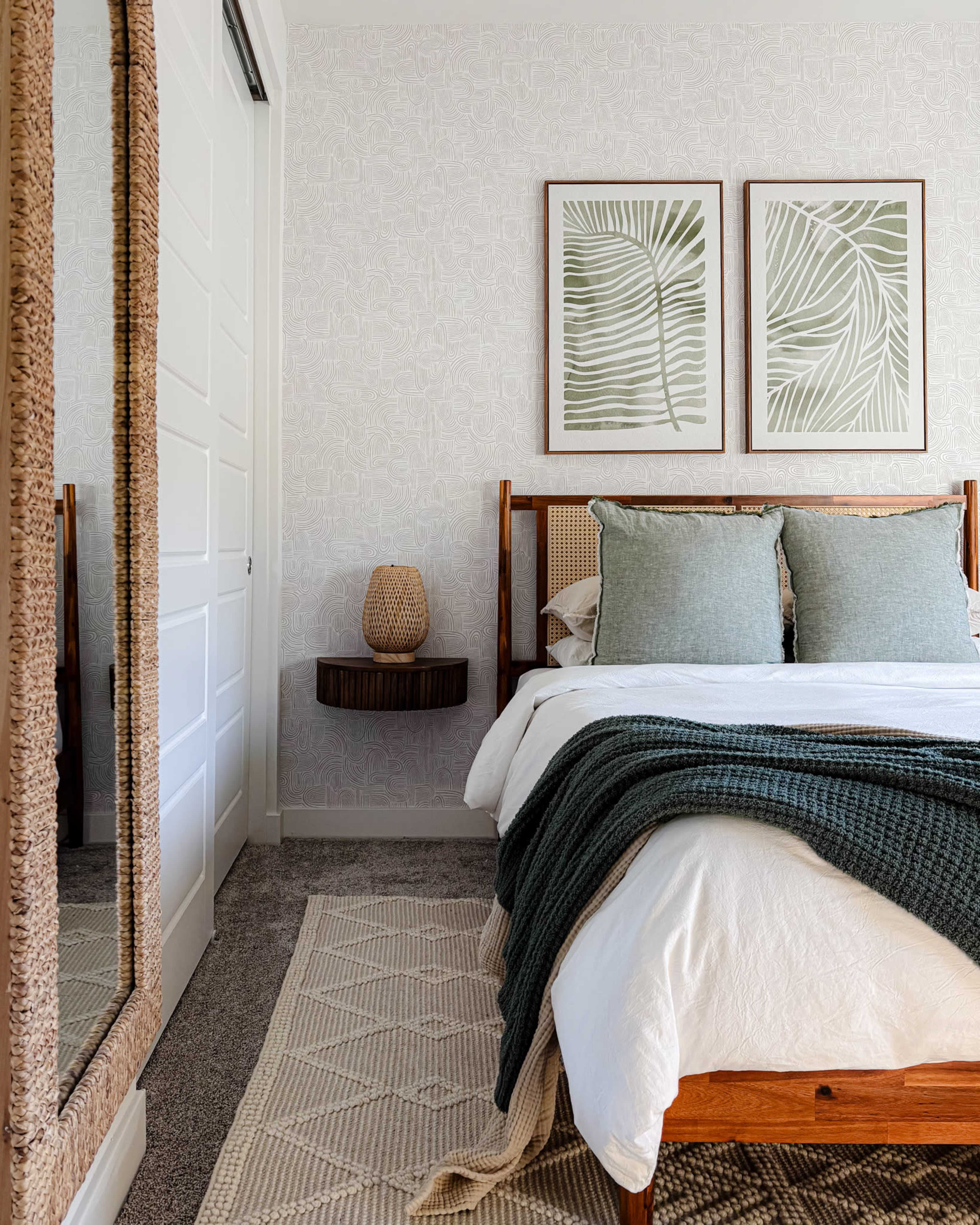 A neatly made bed with green accent pillows and a knit throw is positioned beside a large mirror and botanical wall art in a softly lit bedroom.