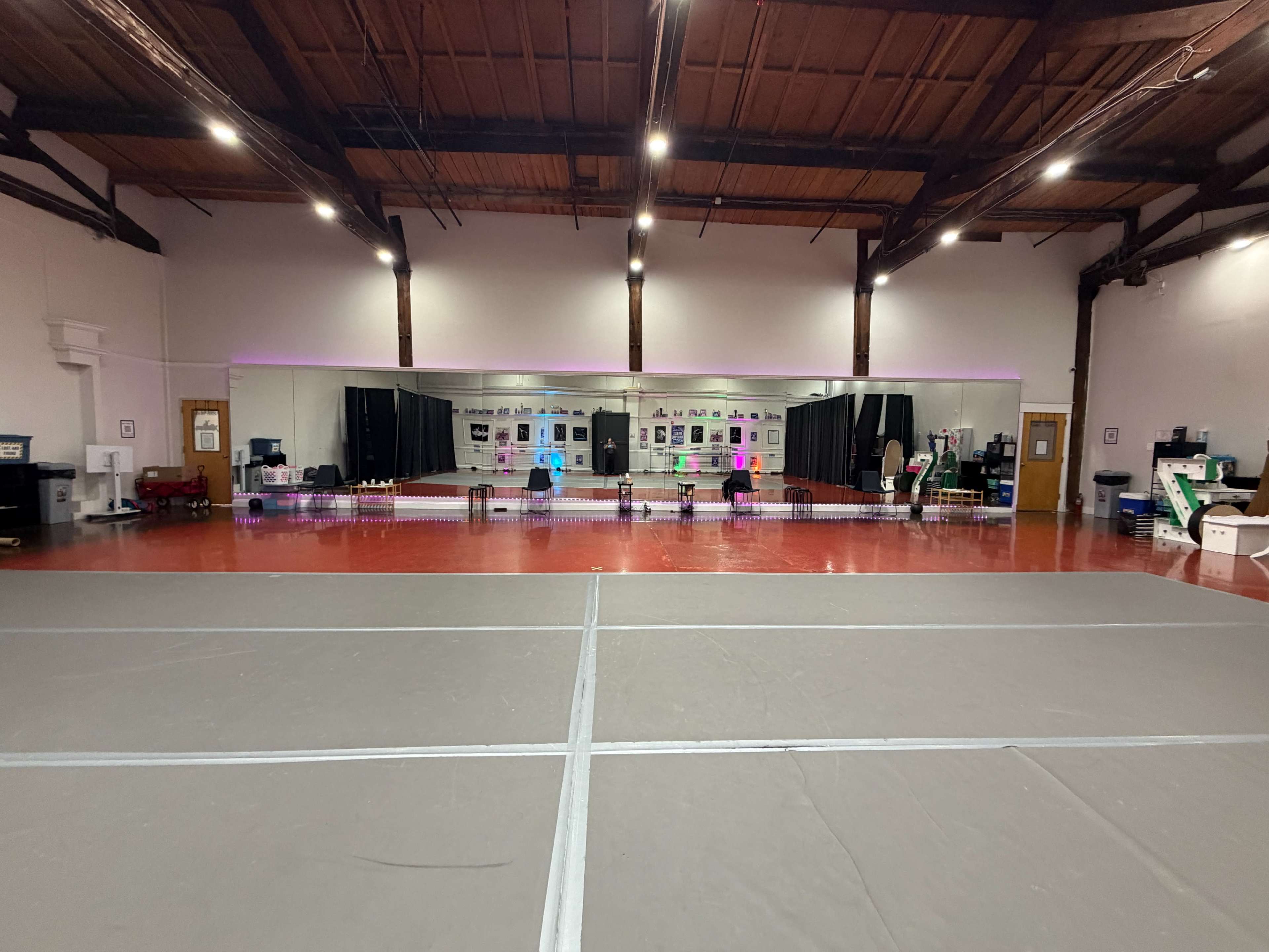 The image shows a spacious dance studio featuring a large mirror, bright lights, and a polished floor, with various equipment and decorations along the back wall.