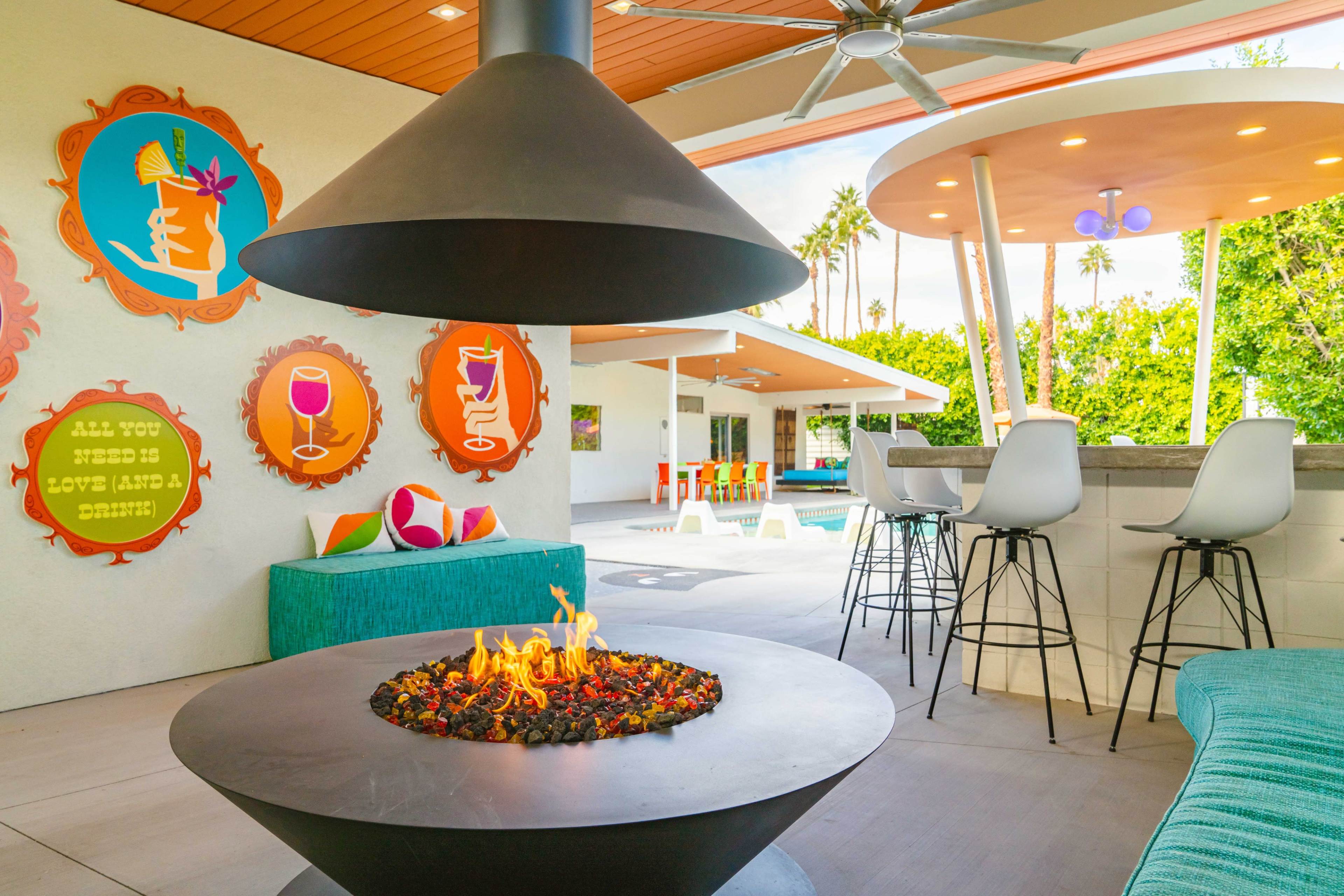 A modern outdoor lounge area features a central fire pit, colorful wall art, and seating near a swimming pool surrounded by palm trees.