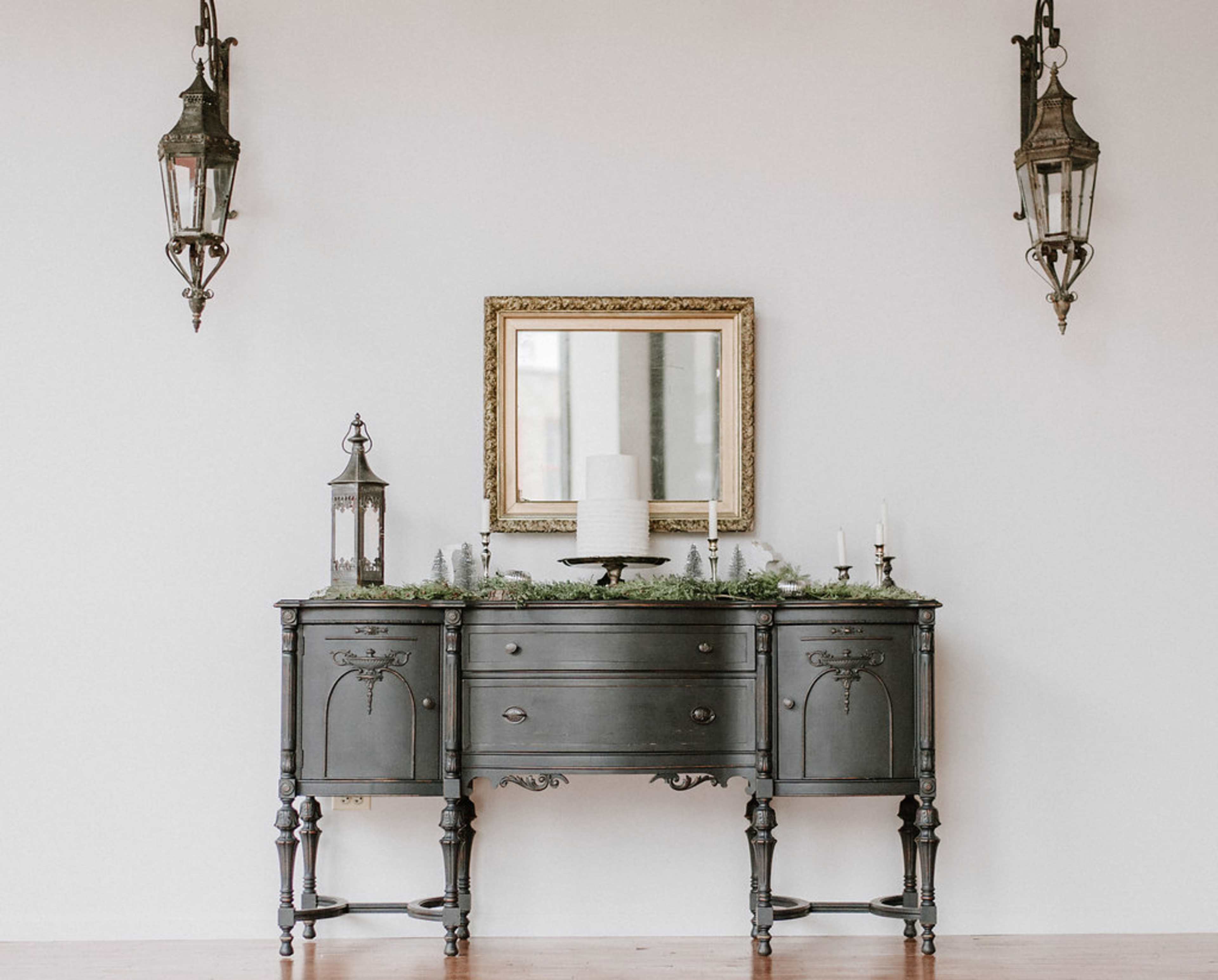 A dark wooden console table with decorative items sits against a white wall, featuring a mirror above and two lanterns flanking the sides.