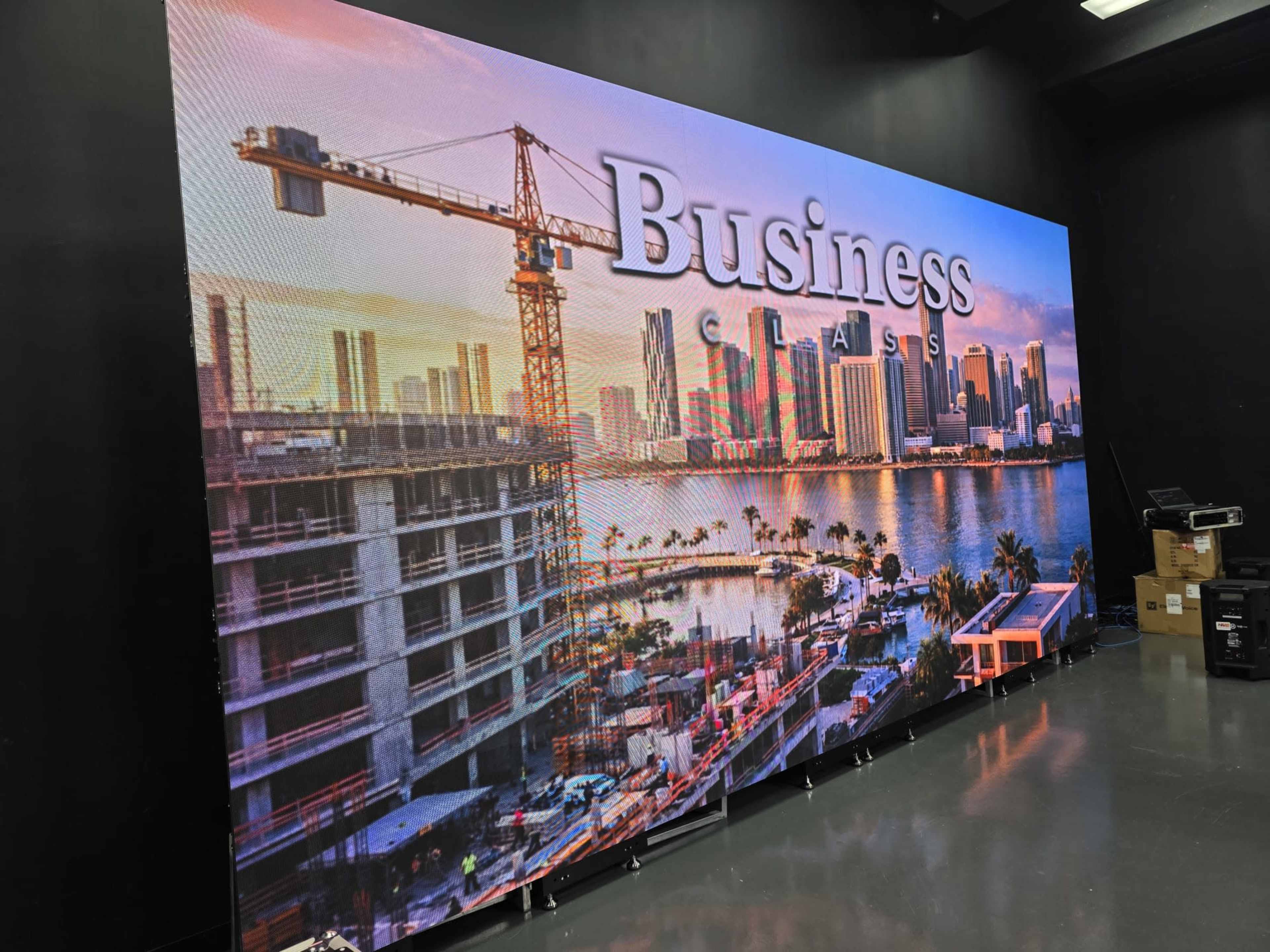 The image features a large digital display showing a construction site with a crane in the foreground and a city skyline across a body of water in the background.