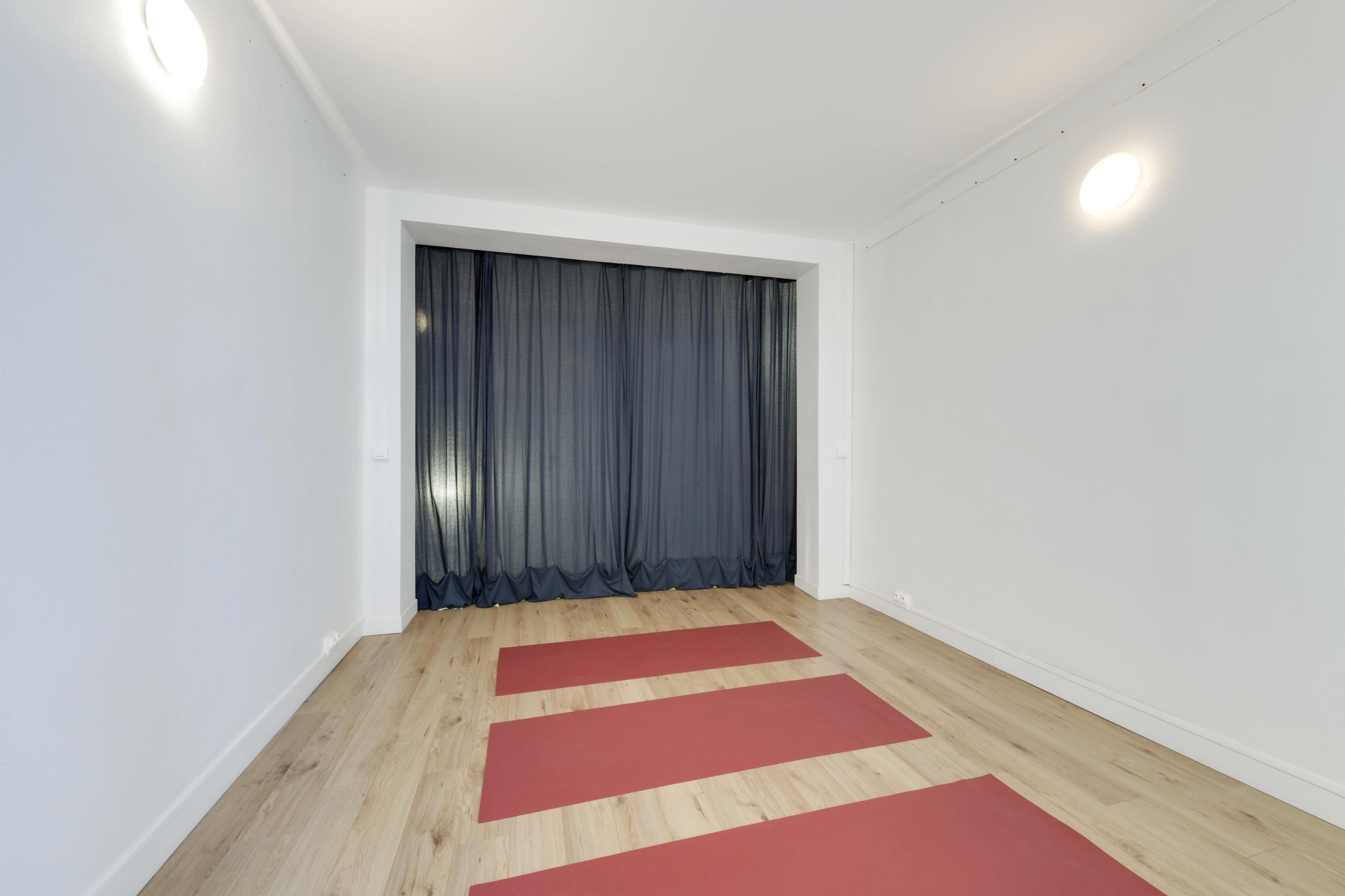 A minimalistic room with wooden flooring features three red yoga mats on the ground and a dark curtain covering a window.