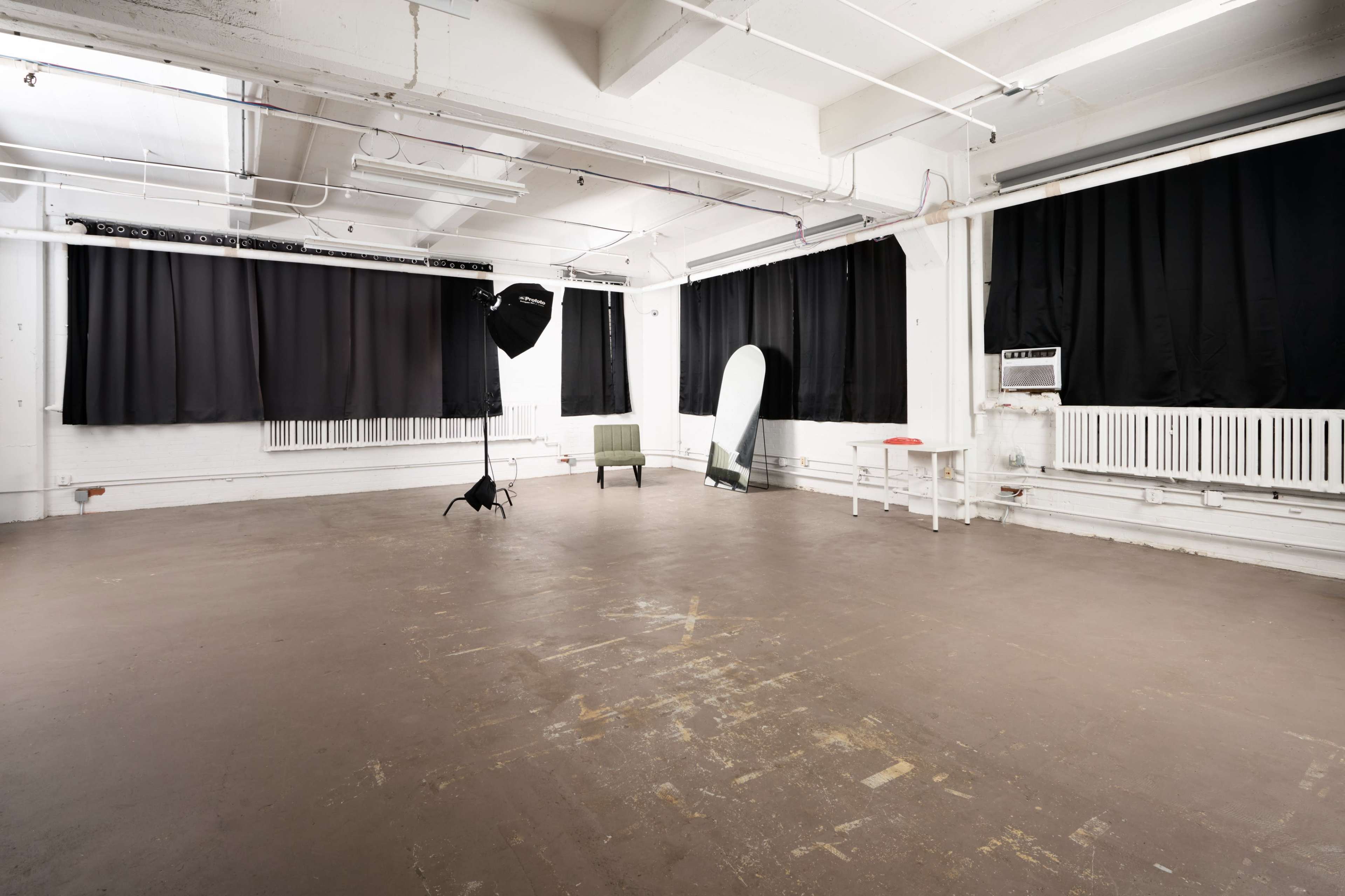 The image shows an empty photo studio with a concrete floor, black curtains, a green chair, a light stand, and a reflector.