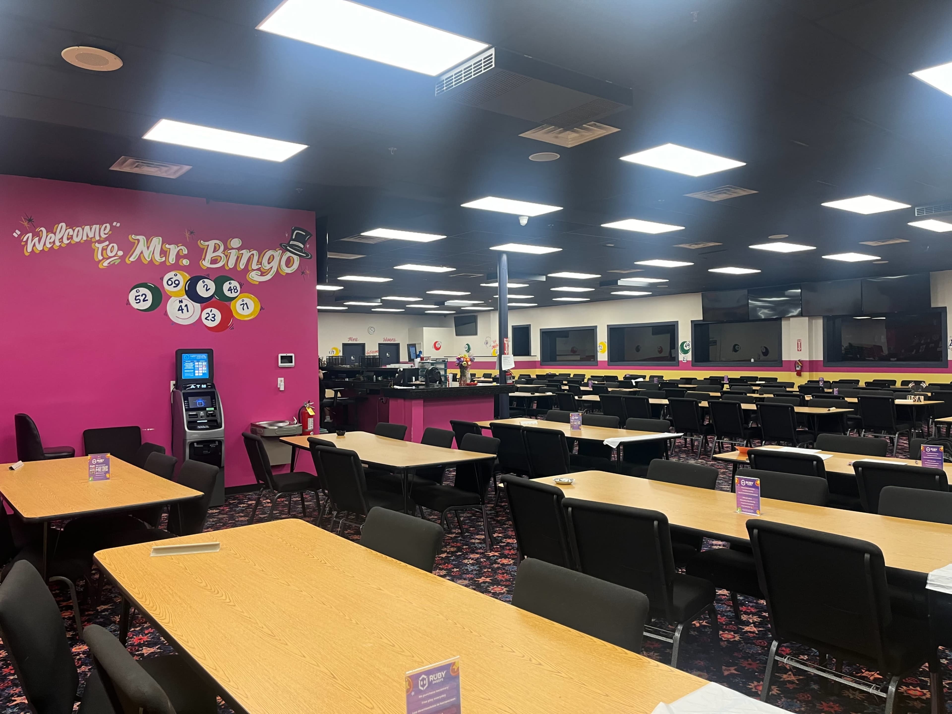 A spacious bingo hall with multiple rows of tables and chairs, brightly lit by overhead lights, and a colorful pink wall featuring the sign "Welcome to Mr. Bingo."