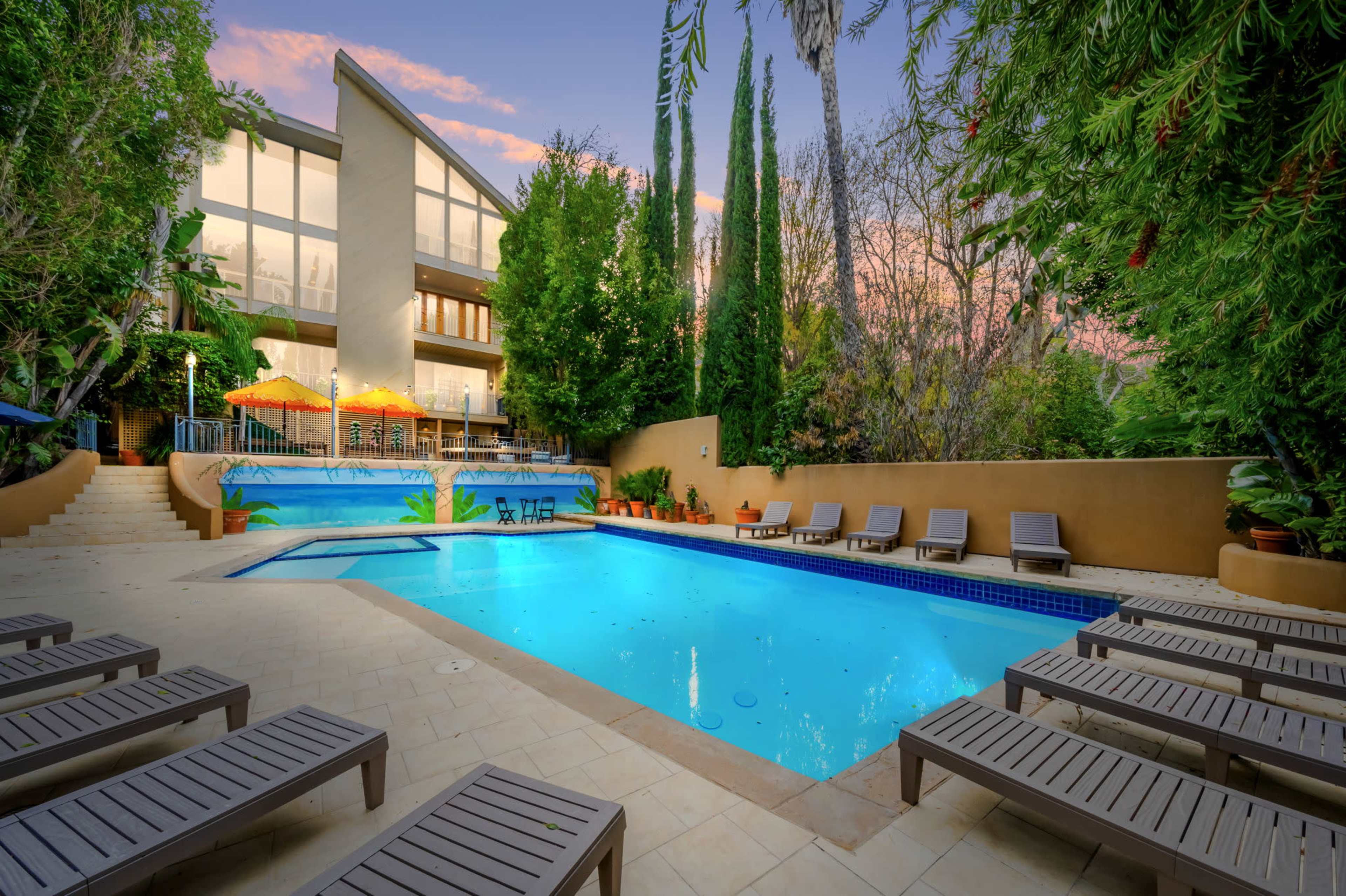 The image shows a modern home with large windows overlooking a swimming pool surrounded by lounge chairs and tropical plants.