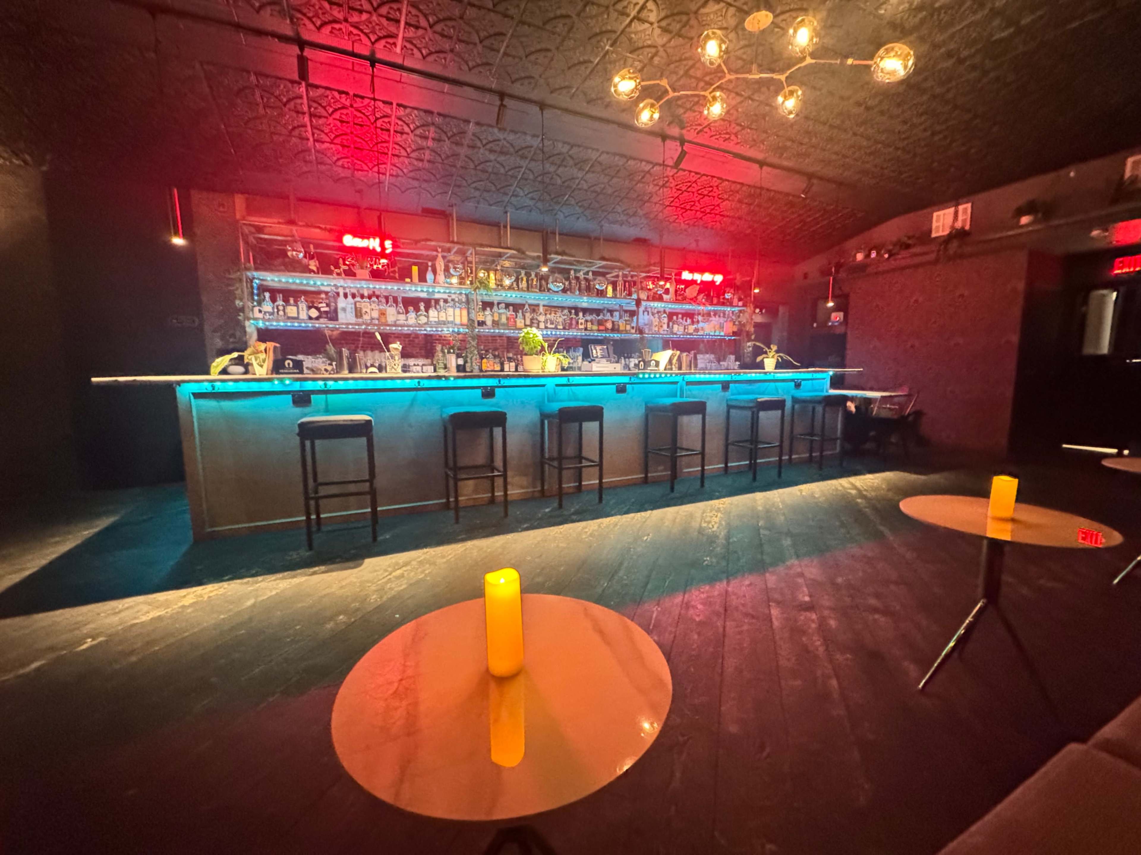 The interior of a dimly lit bar features a glowing blue backlit bar with stools and illuminated shelves filled with bottles, accompanied by round tables with candles casting a warm glow.
