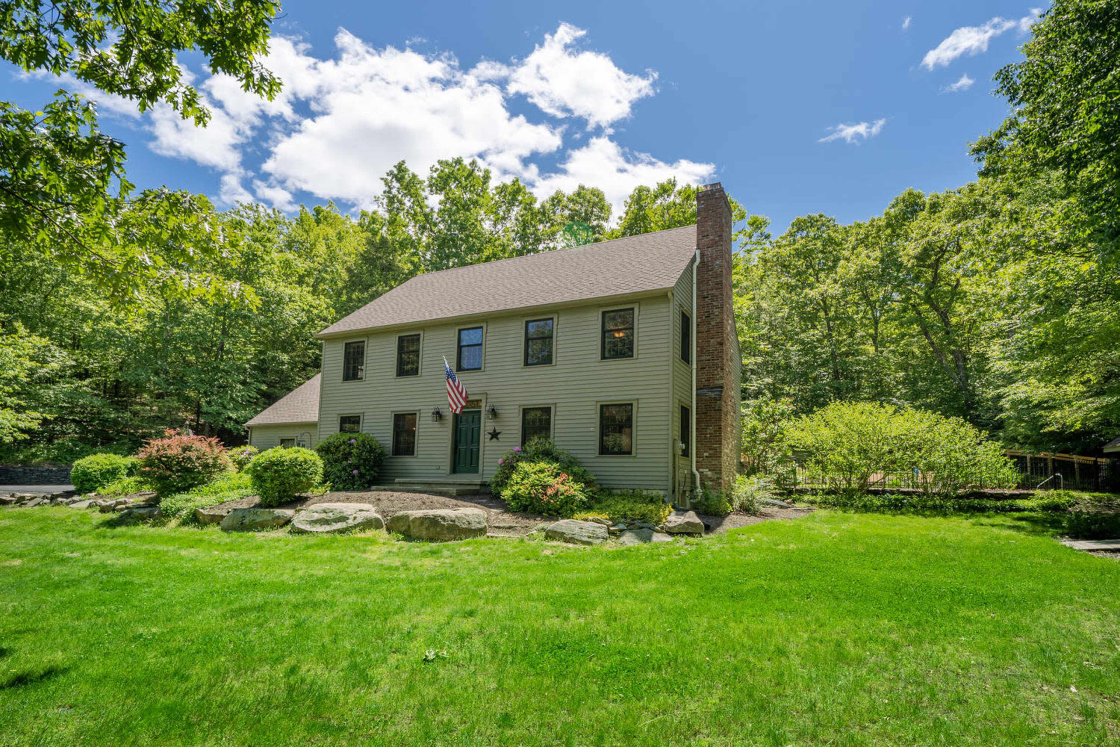 A two-story green house with a chimney and an American flag stands on a grassy lawn surrounded by trees and landscaped stones.
