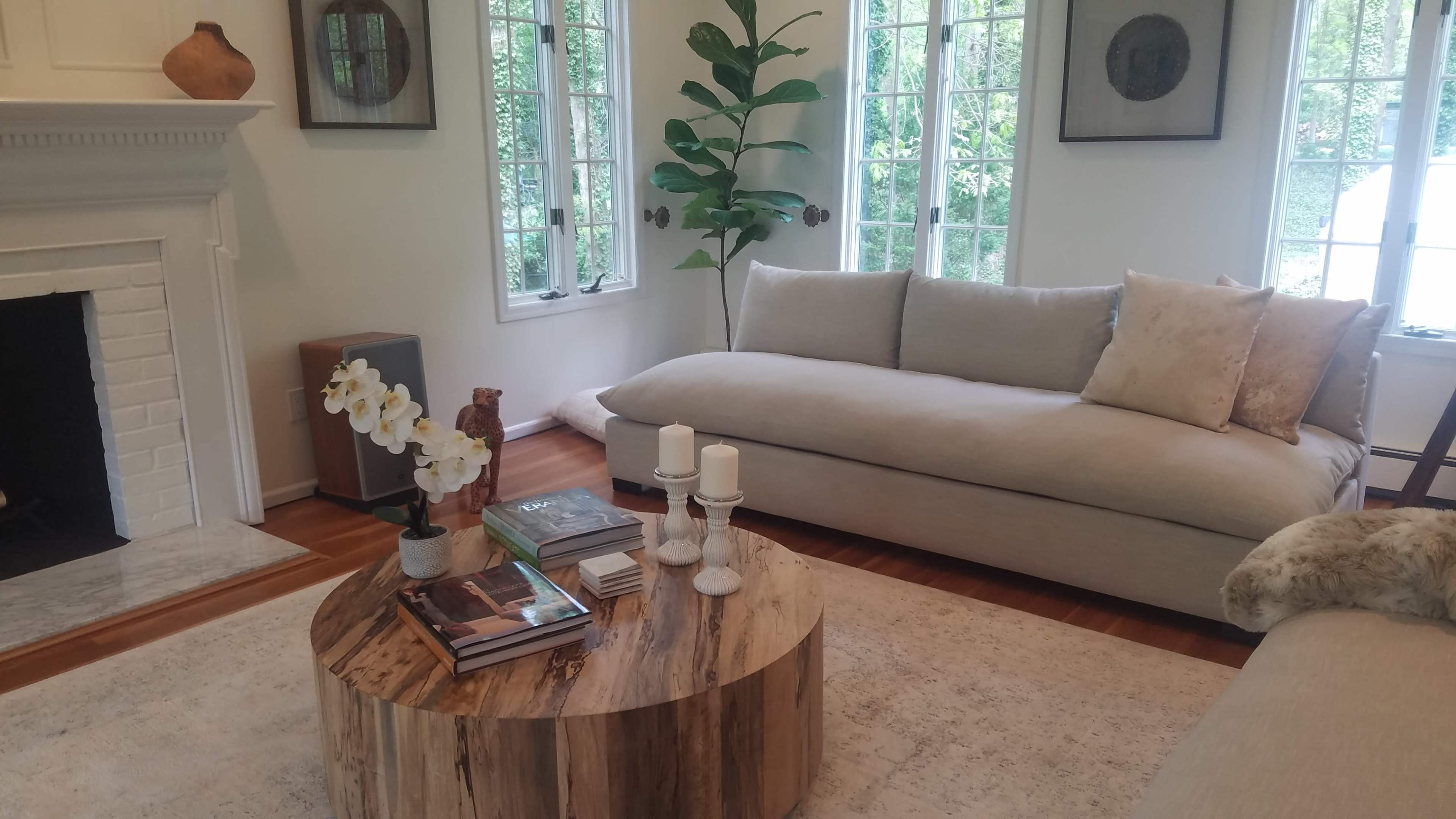 The image shows a modern living room with a light-colored sofa, a round wooden coffee table, and large windows that allow natural light to enter the space.