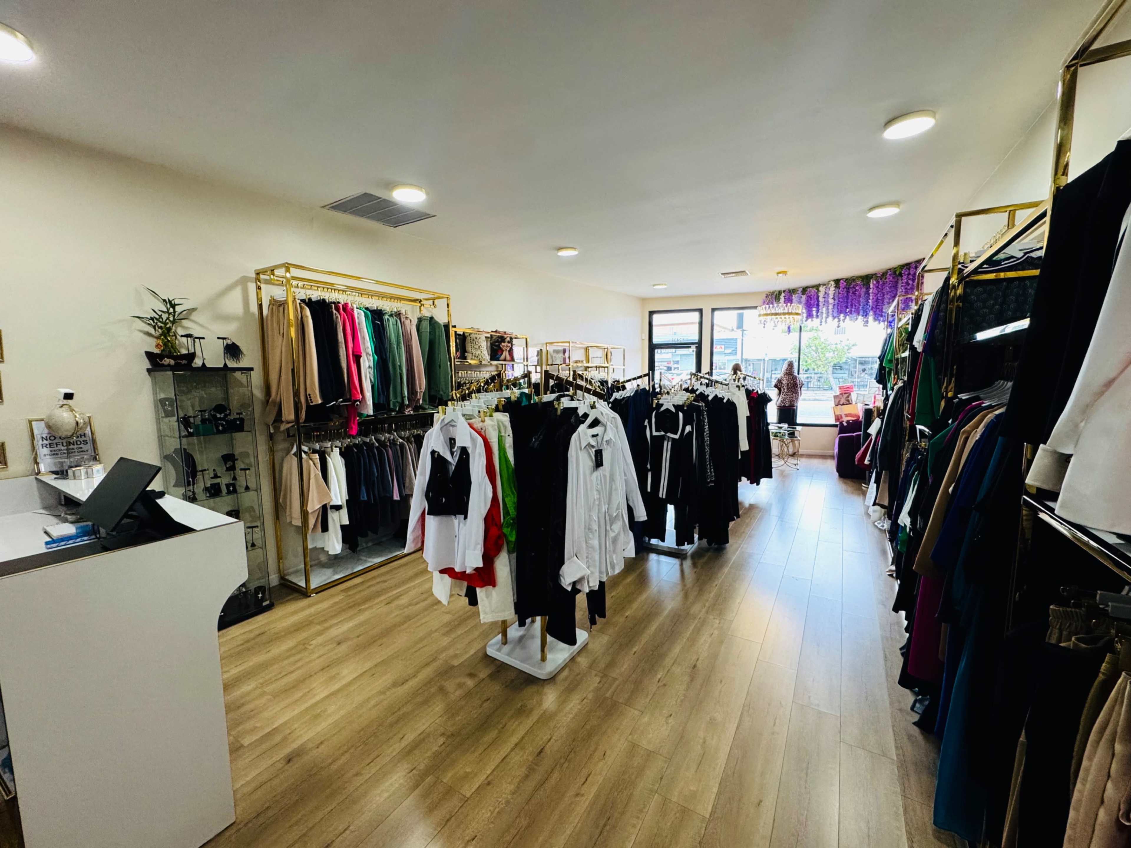 The image shows an interior view of a clothing store with racks of garments organized along the walls and a checkout counter in the foreground.
