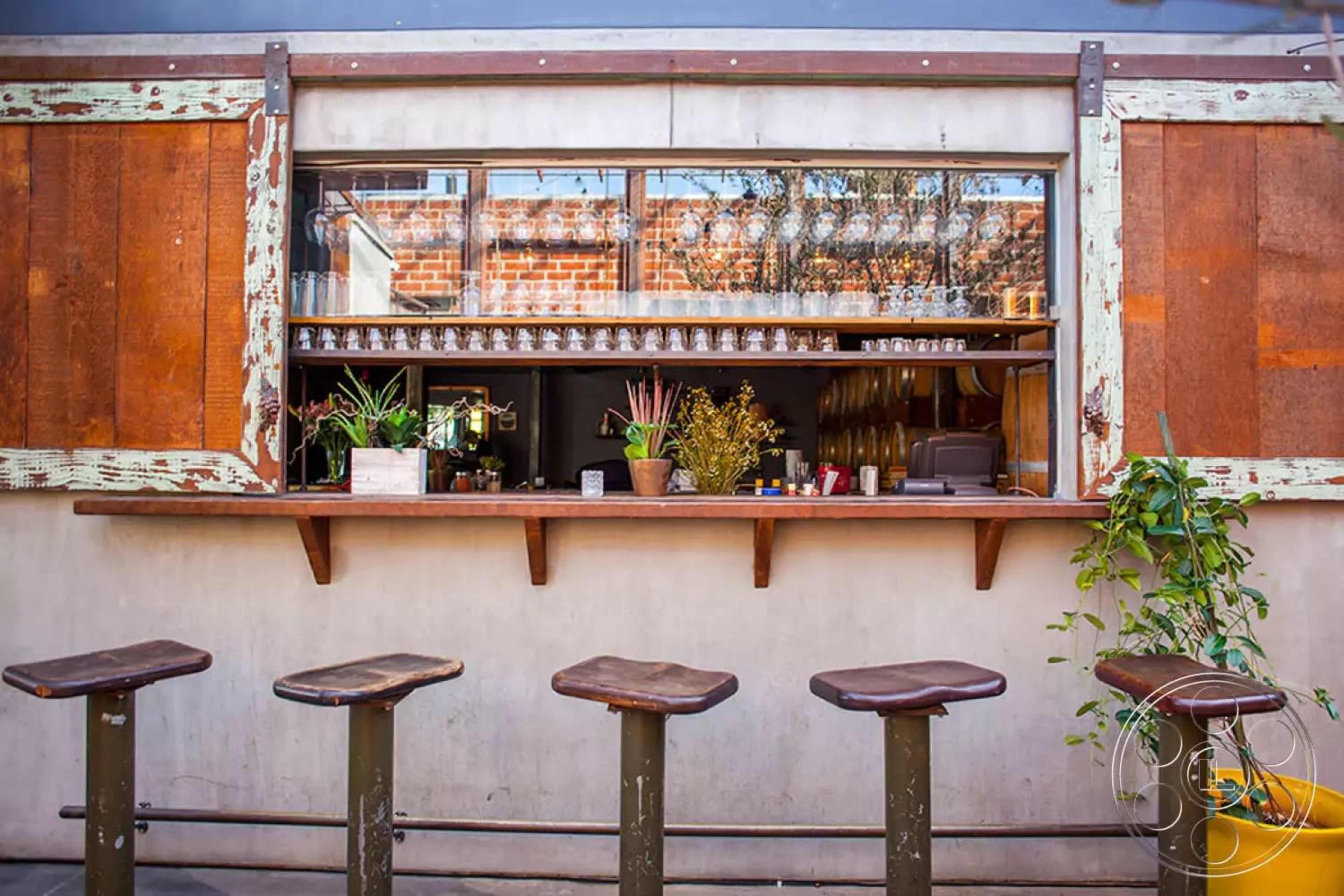 The image shows a rustic bar counter with wooden stools arranged in front of a window displaying various glassware and plants.