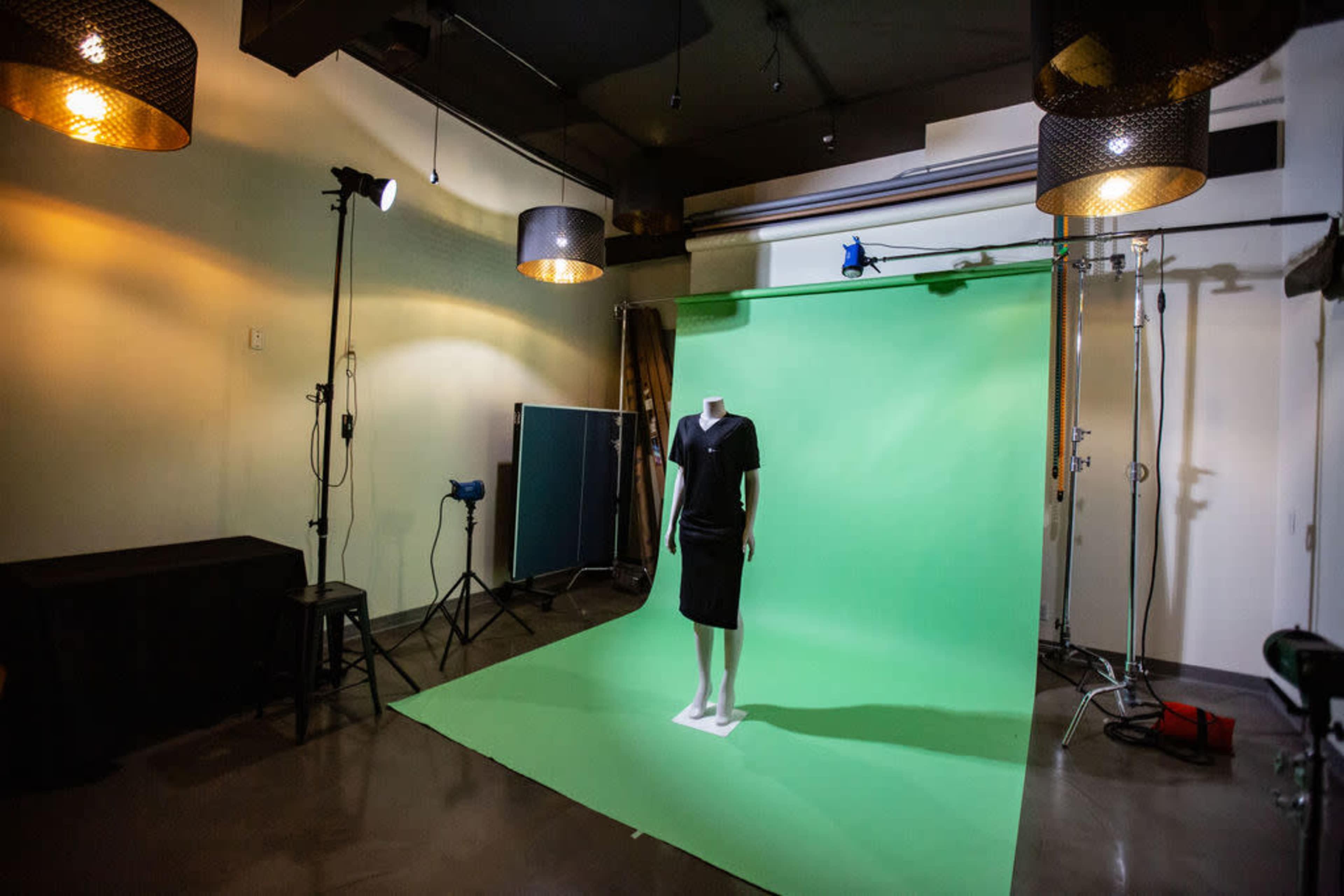 A black dress is displayed on a mannequin in a photography studio with a green backdrop and studio lighting equipment.