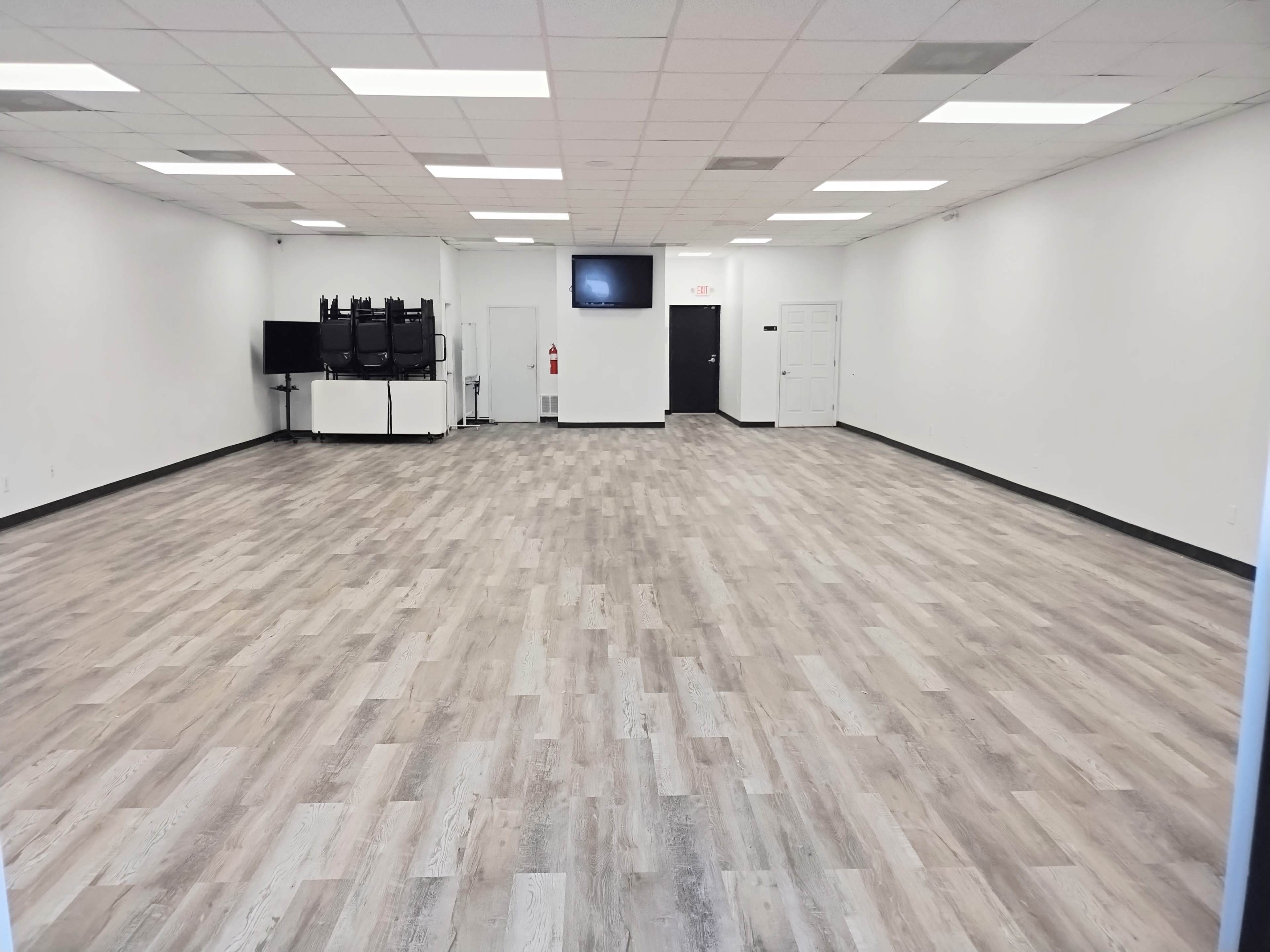 A large, empty room with light wood flooring, white walls, and a few pieces of furniture arranged near the back.