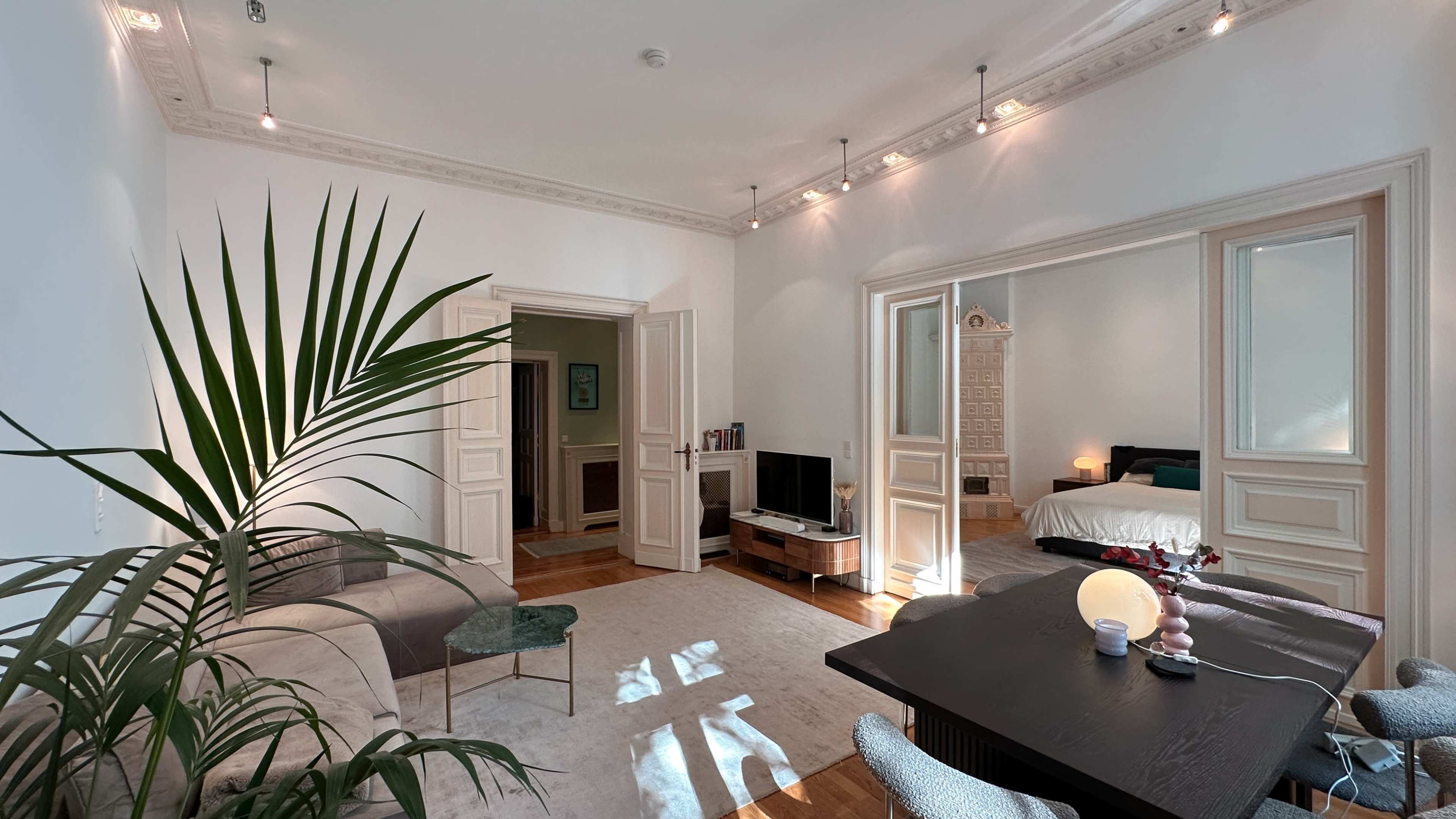 The image shows a bright living space featuring a large plant, a sofa, a dining table, and a bedroom visible through an open doorway.