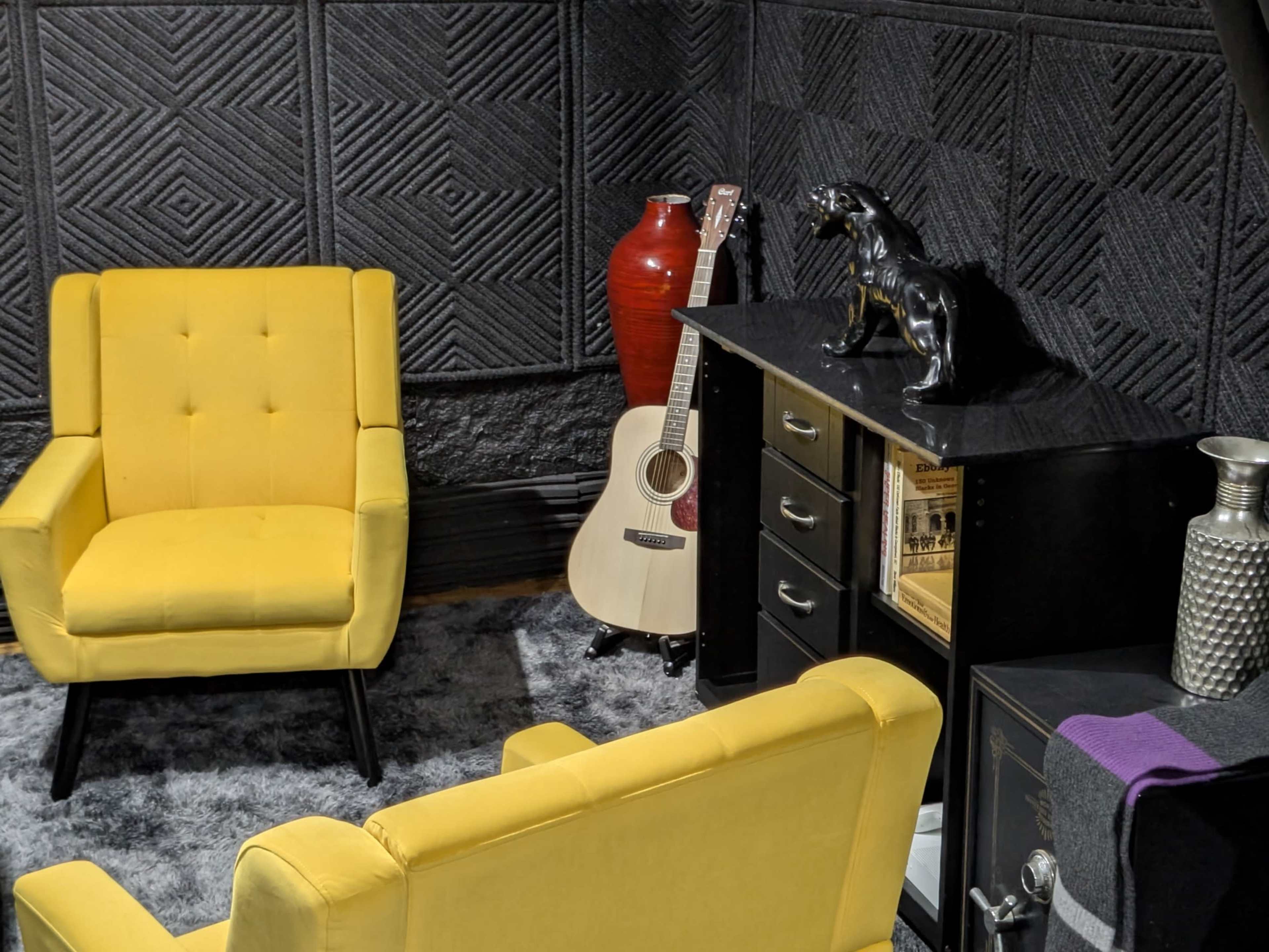The image features two yellow armchairs facing a black cabinet with a guitar and decorative items in a cozy room with textured walls.